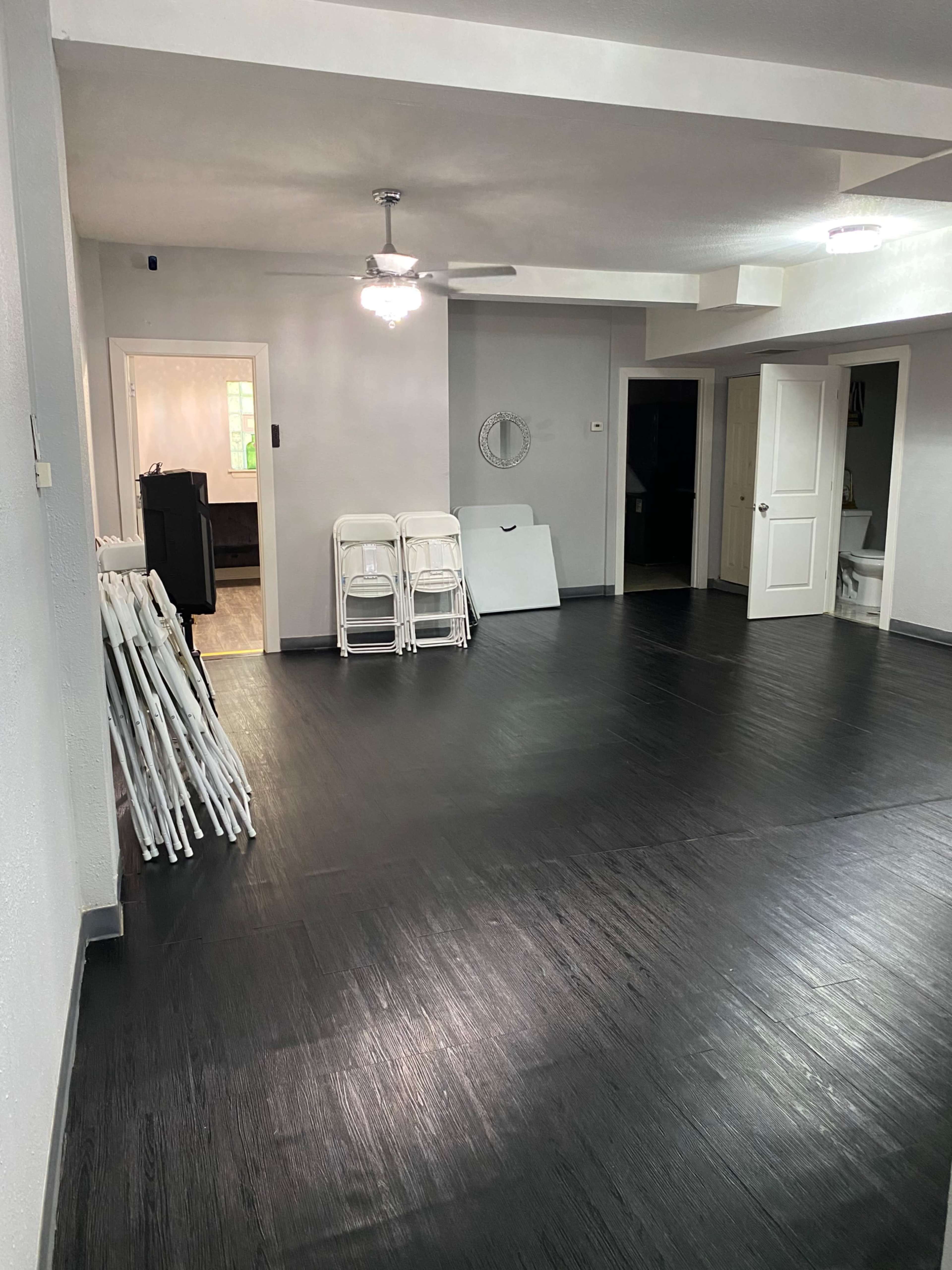 The image shows a spacious, empty room with black flooring, a ceiling fan, and a few white folding chairs stacked against the wall.