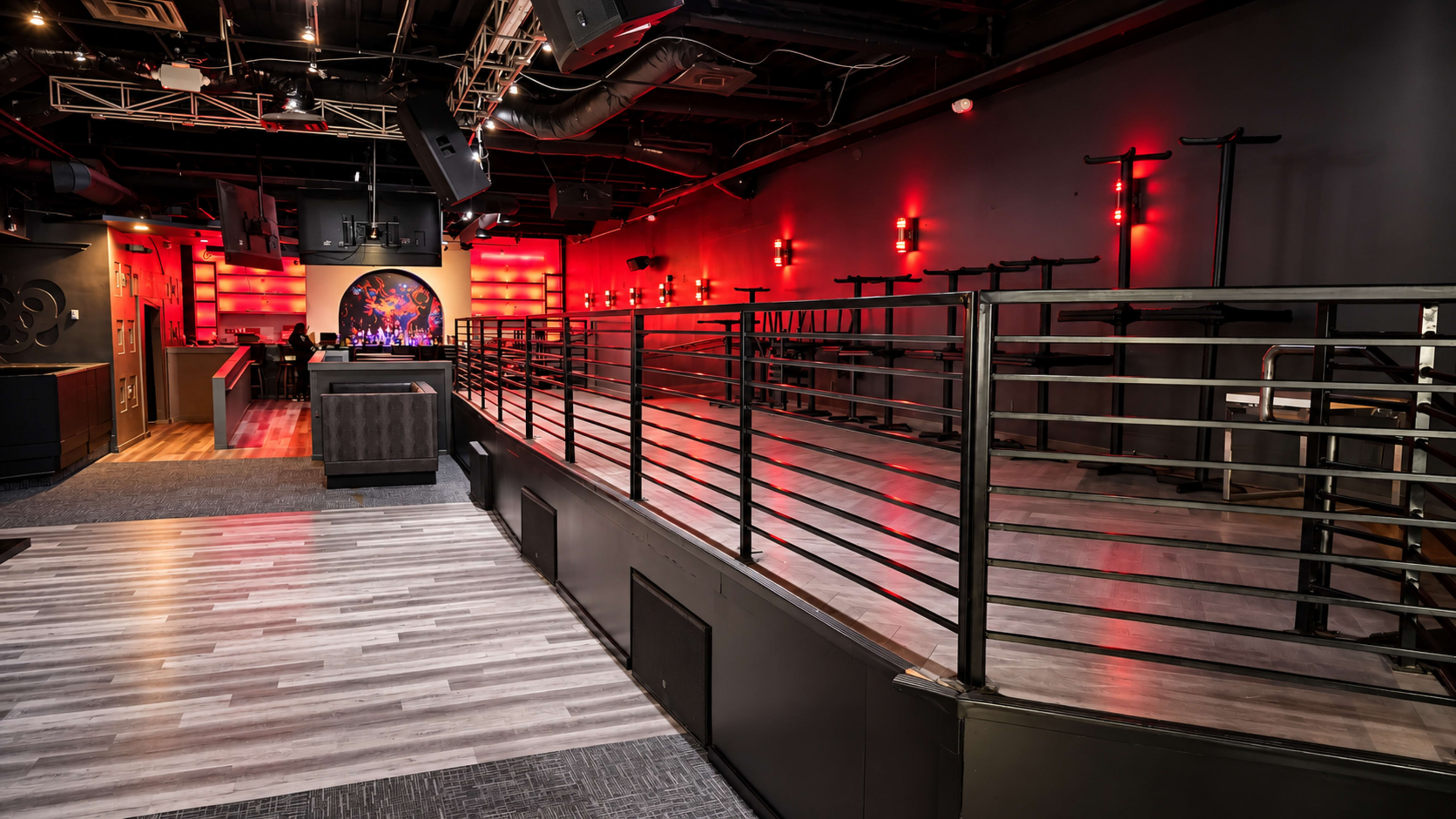 The image shows an empty music venue with a stage at the front, featuring red lighting and a raised area with a railing.