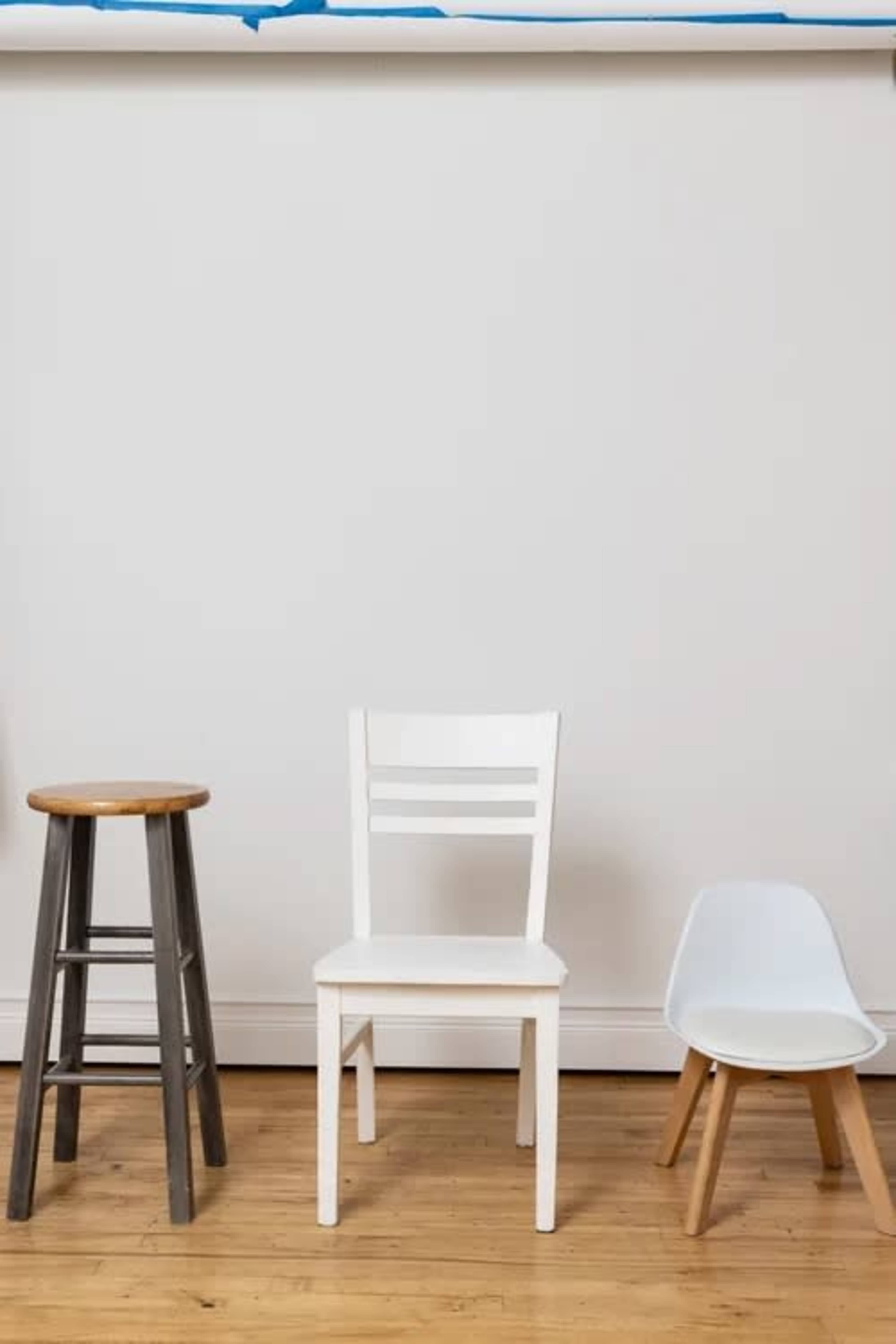 The image shows three chairs of different designs positioned against a plain white wall on a wooden floor.