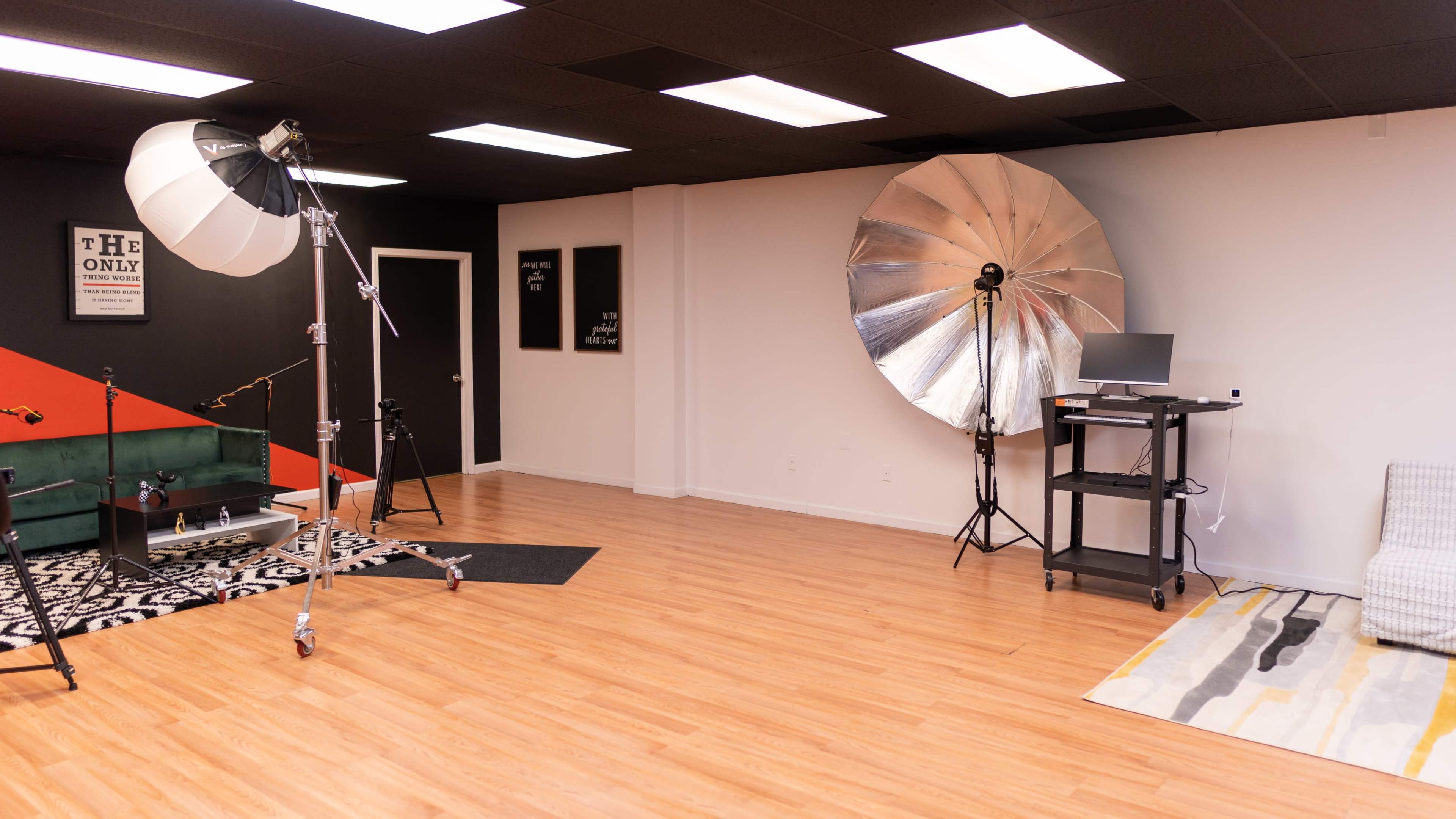 A spacious photography studio featuring a large silver reflector, a softbox light, a computer station, and various props arranged on a wooden floor.