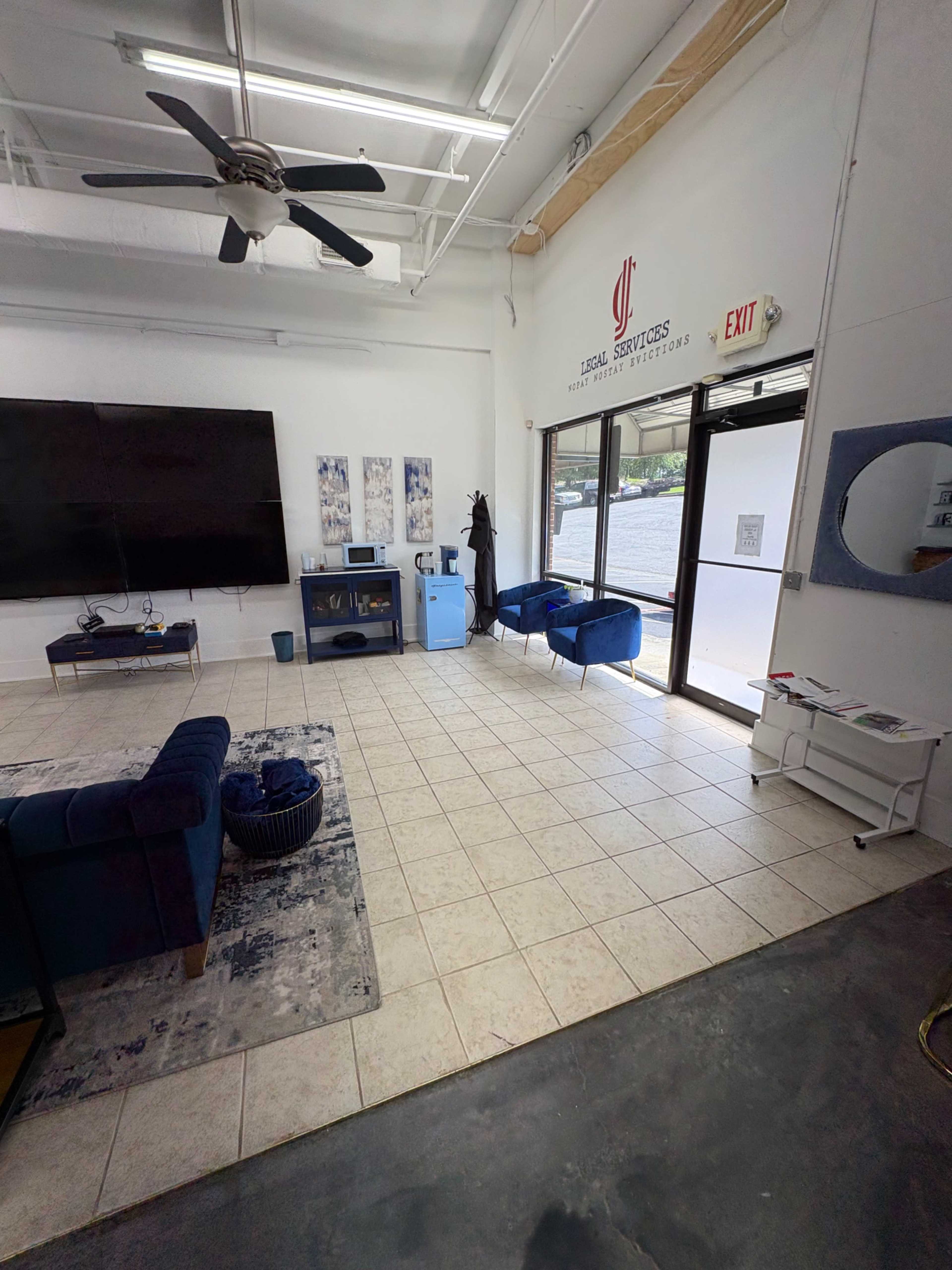 The image shows a well-lit waiting area with blue sofas, a large television on the wall, and an exit door leading outside.