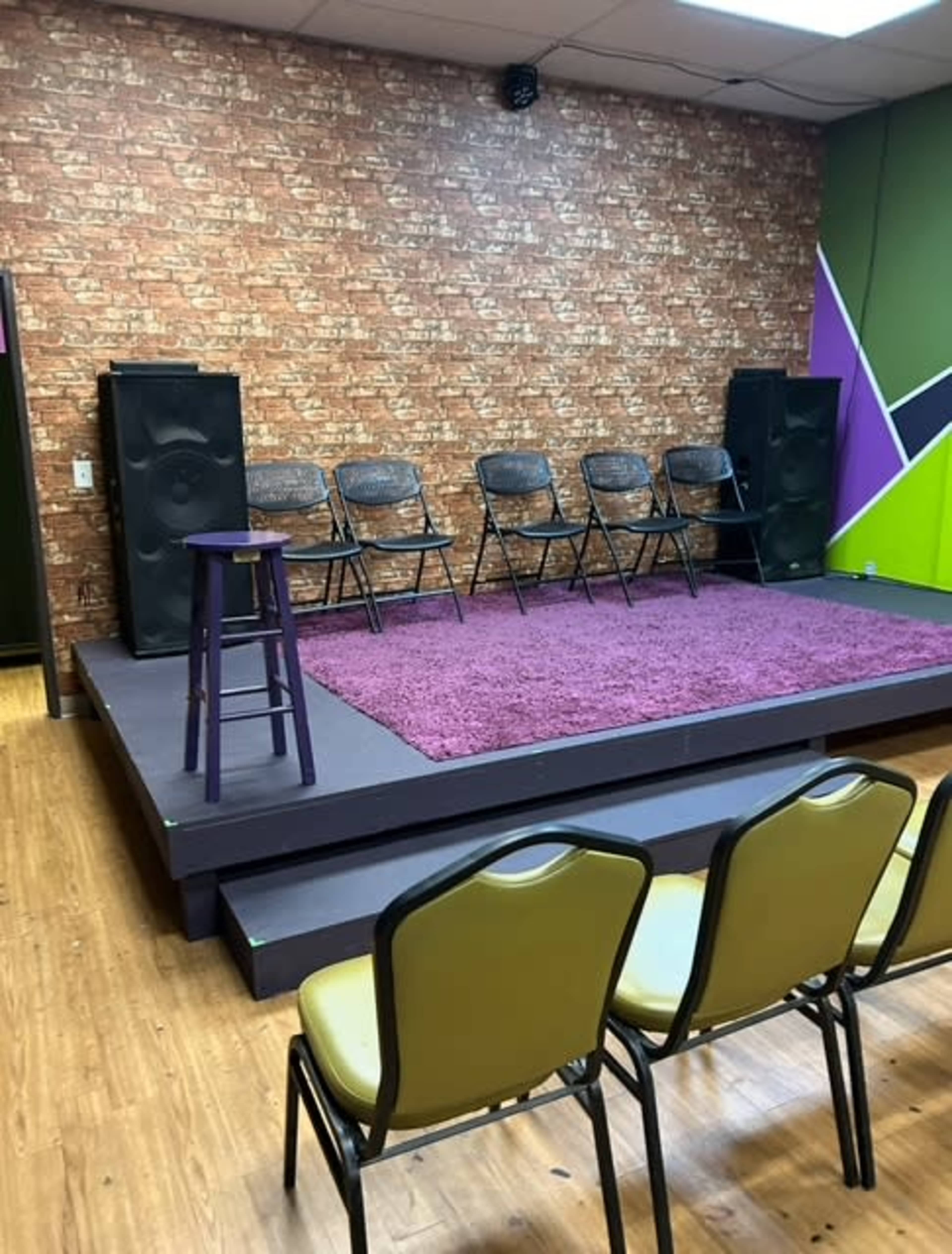 The image shows a small stage with five black folding chairs set up on it, surrounded by a purple rug, and facing an audience area with yellow chairs.