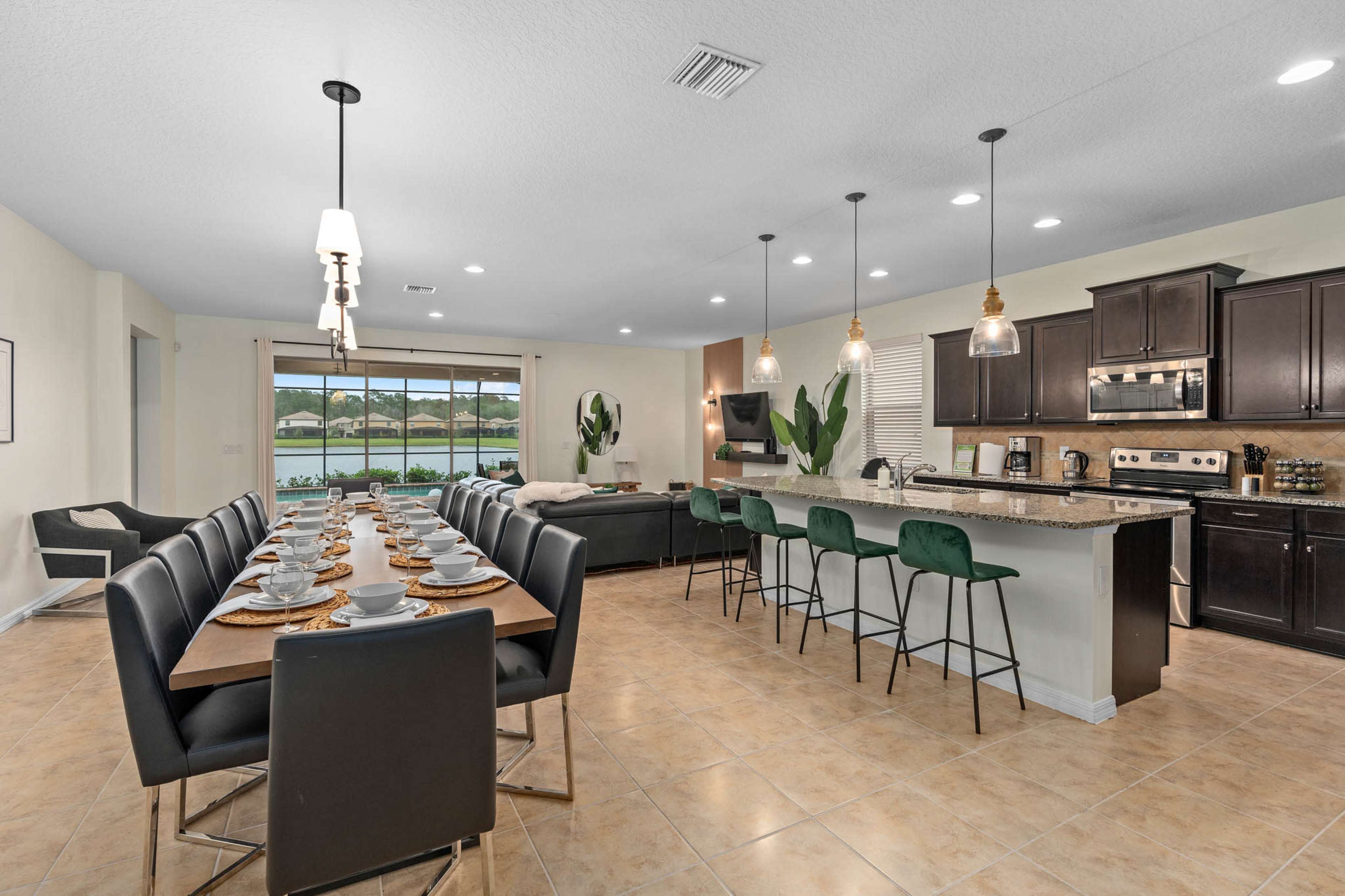 The image shows a spacious dining area with a long table set for a meal, adjacent to a modern kitchen with a large island and bar stools.