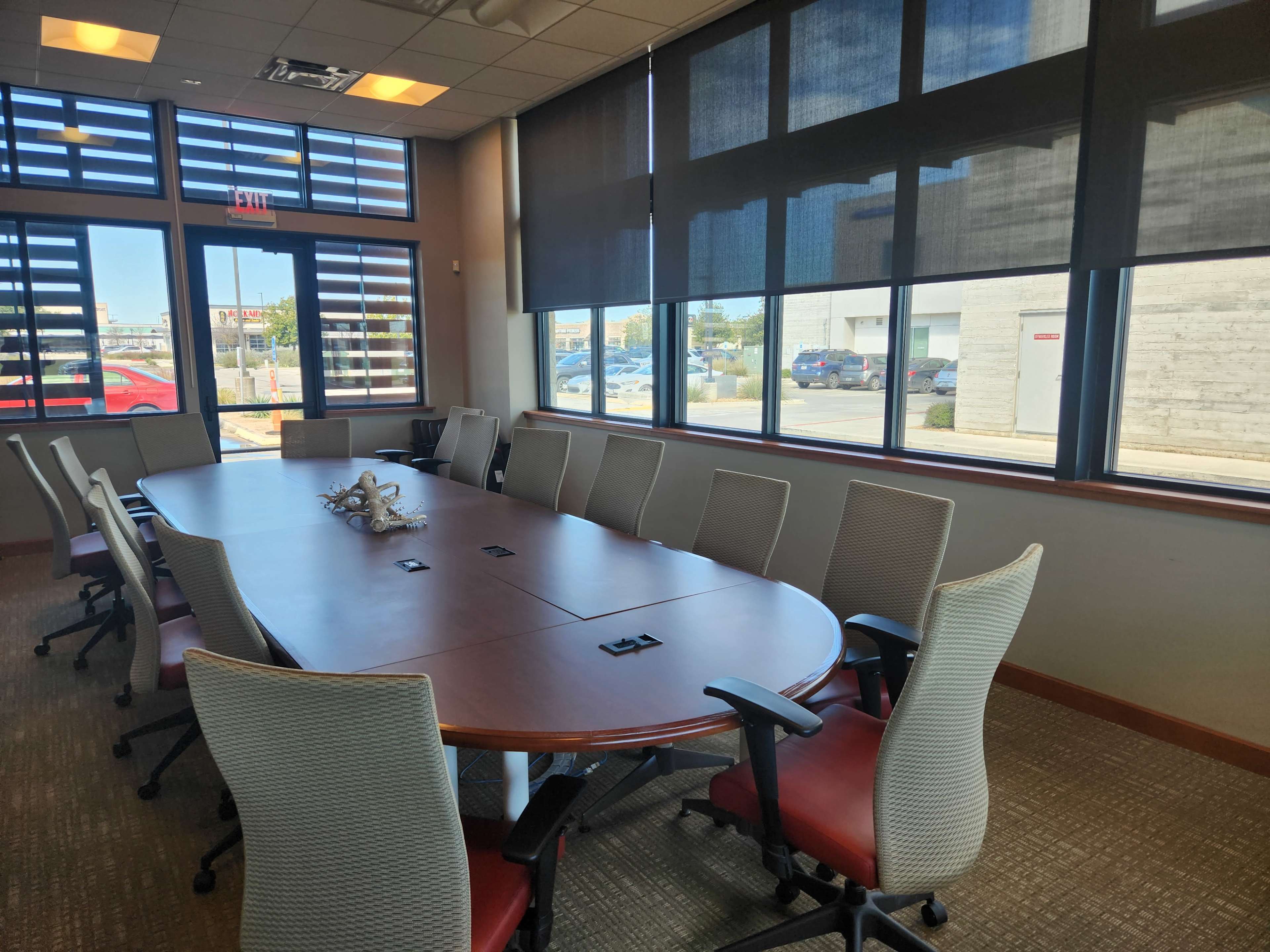 A large, oval conference table with white chairs is set up in a well-lit meeting room featuring large windows and blinds.