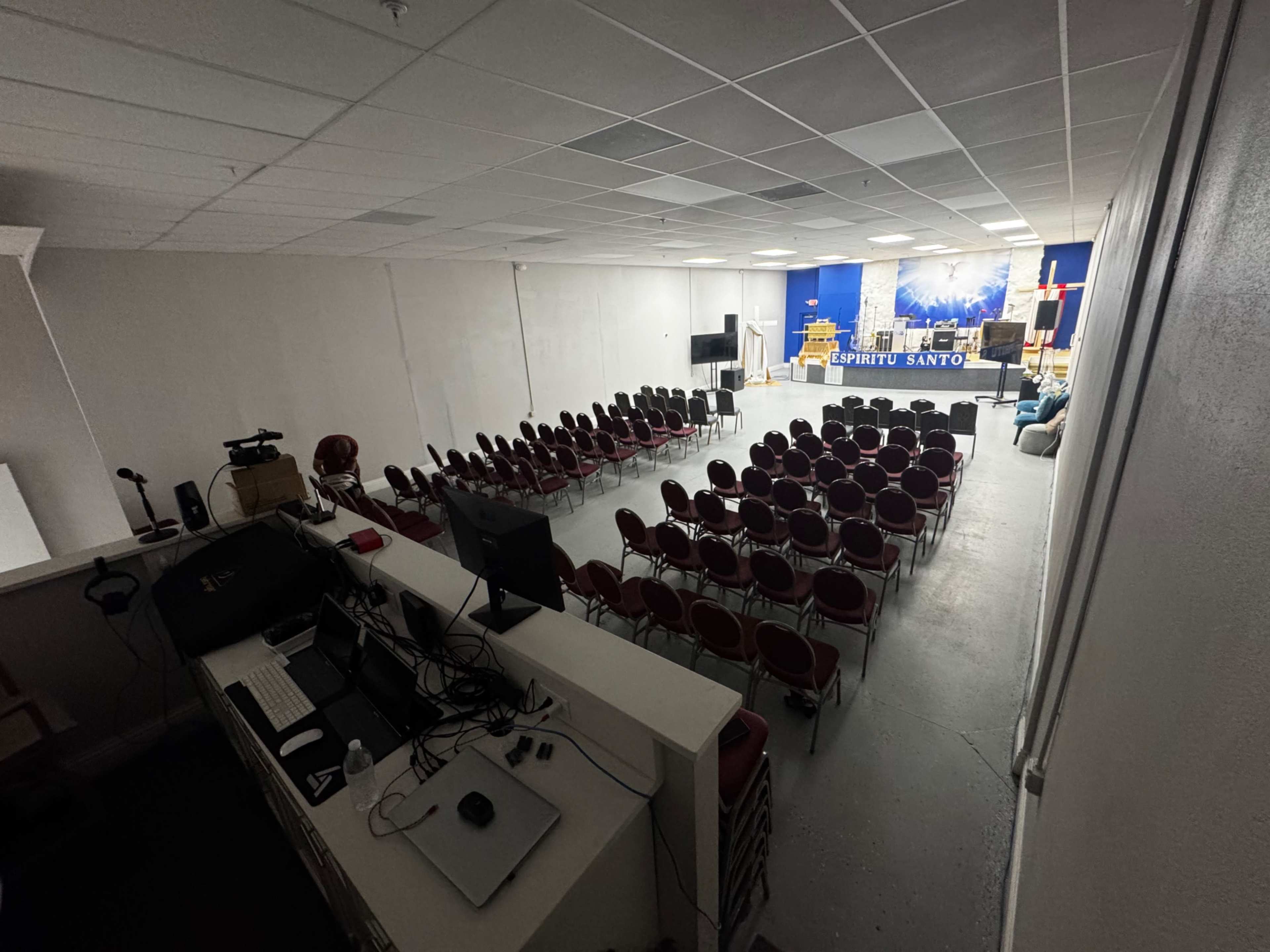 The image shows an empty auditorium with rows of red chairs facing a stage that features a blue backdrop and audio equipment.