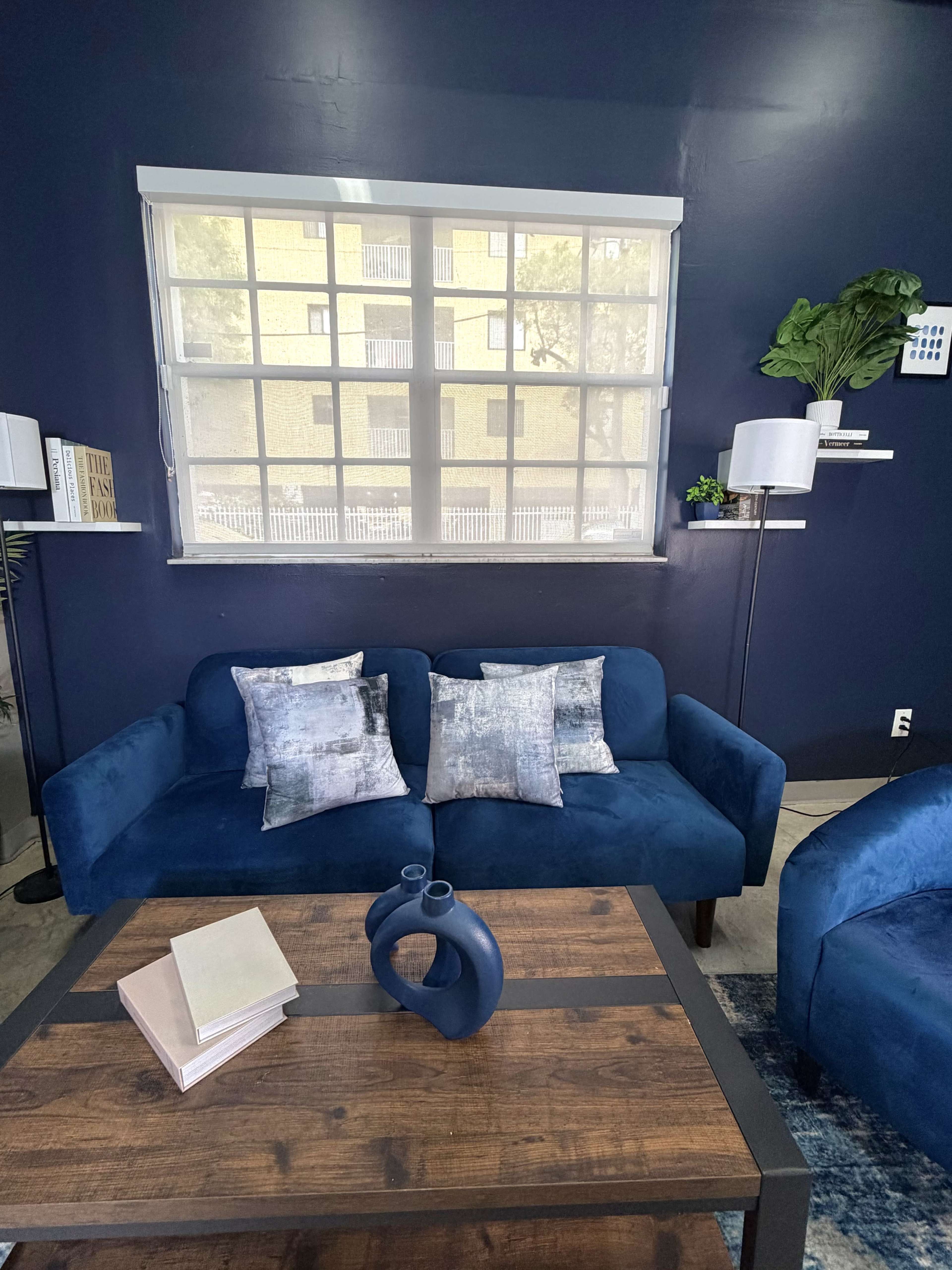 The image shows a modern living room setup with a blue couch, two decorative pillows, a coffee table with a book and a vase, and a large window in the background.