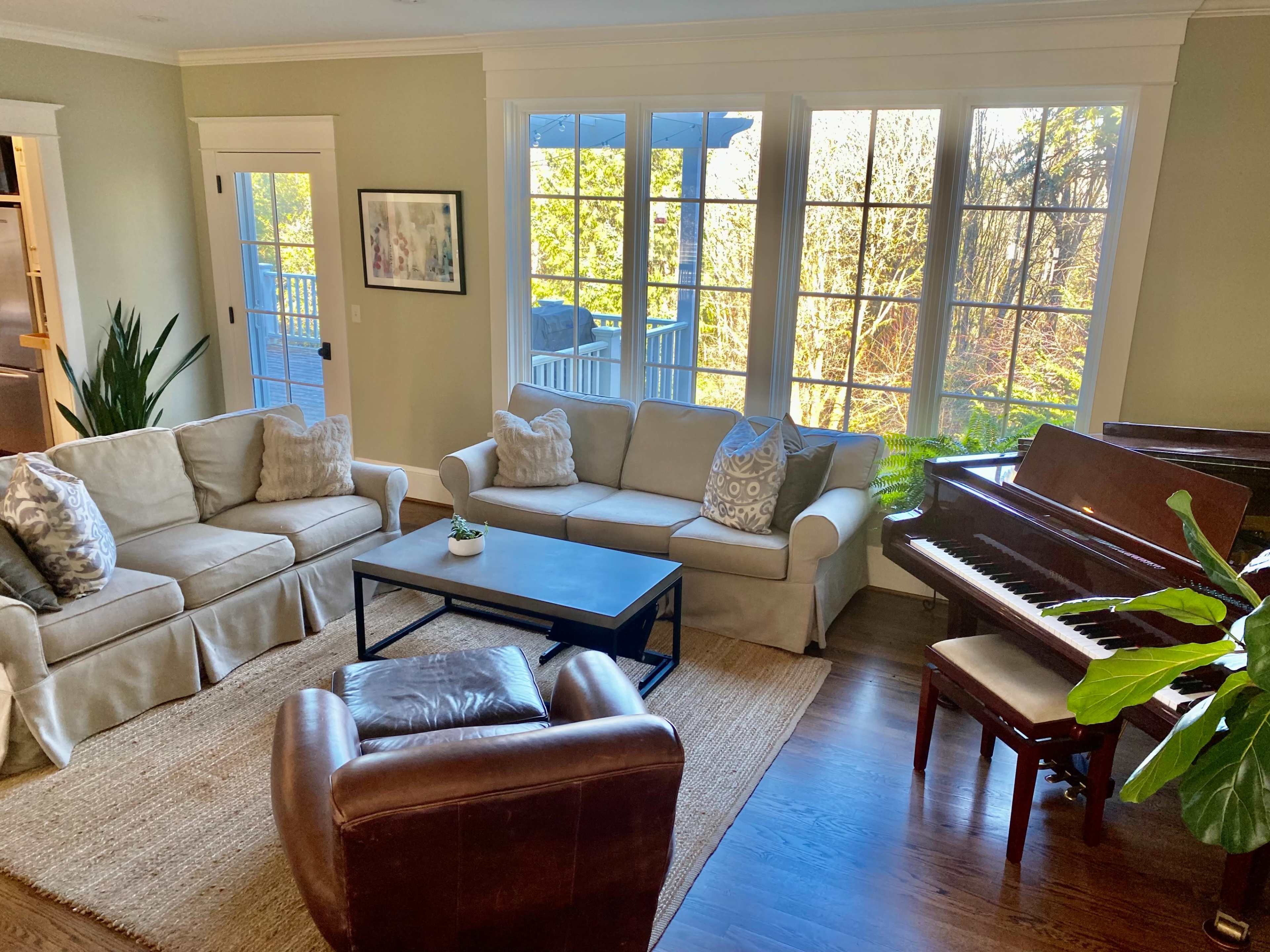 A cozy living room features two sofas, a coffee table, a leather armchair, and a piano, with large windows providing a view of greenery outside.