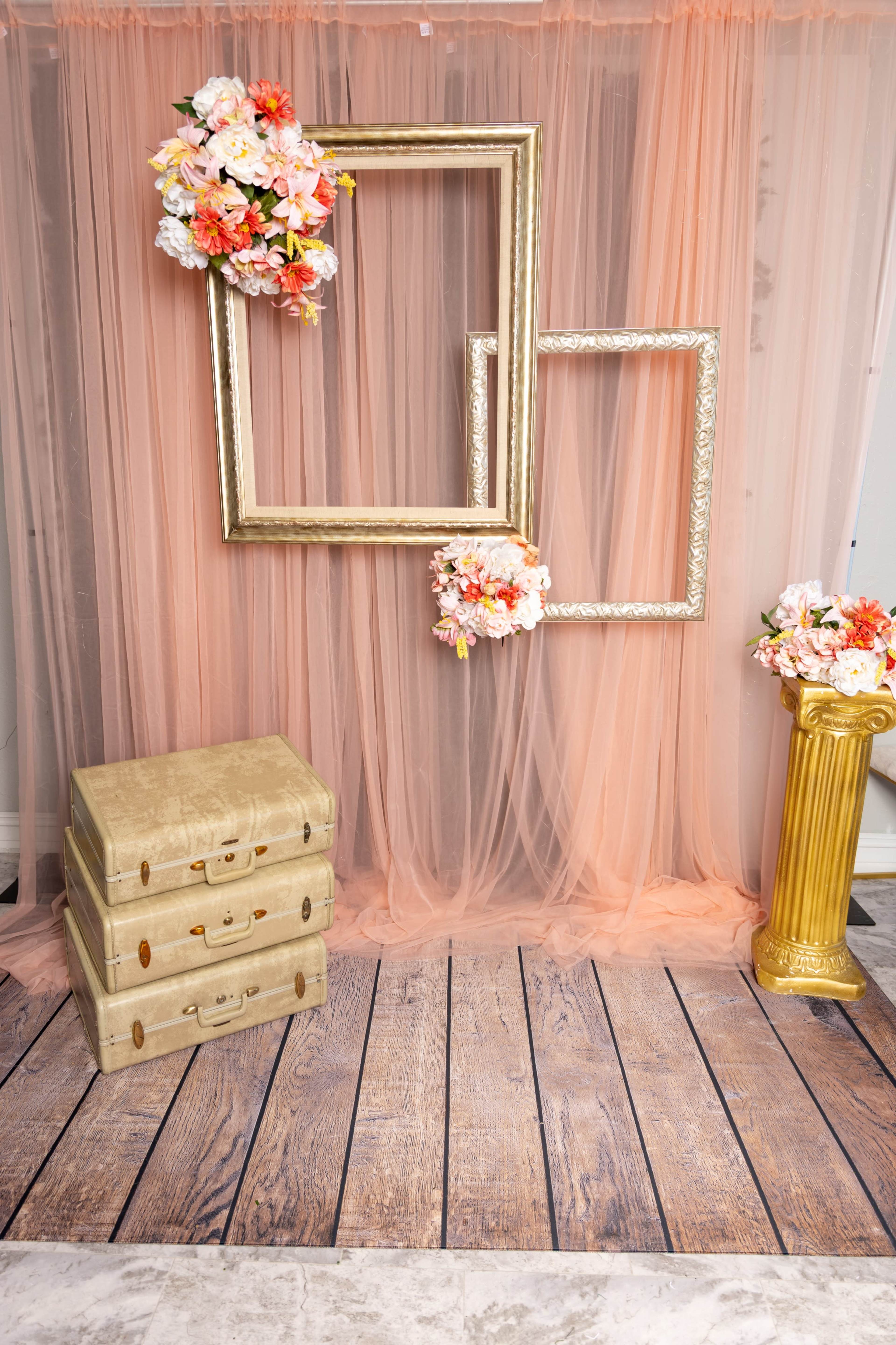 The scene features a peach-colored backdrop with three empty ornate frames, two floral arrangements, and a golden column, complemented by stacked vintage-style suitcases on a wooden floor.