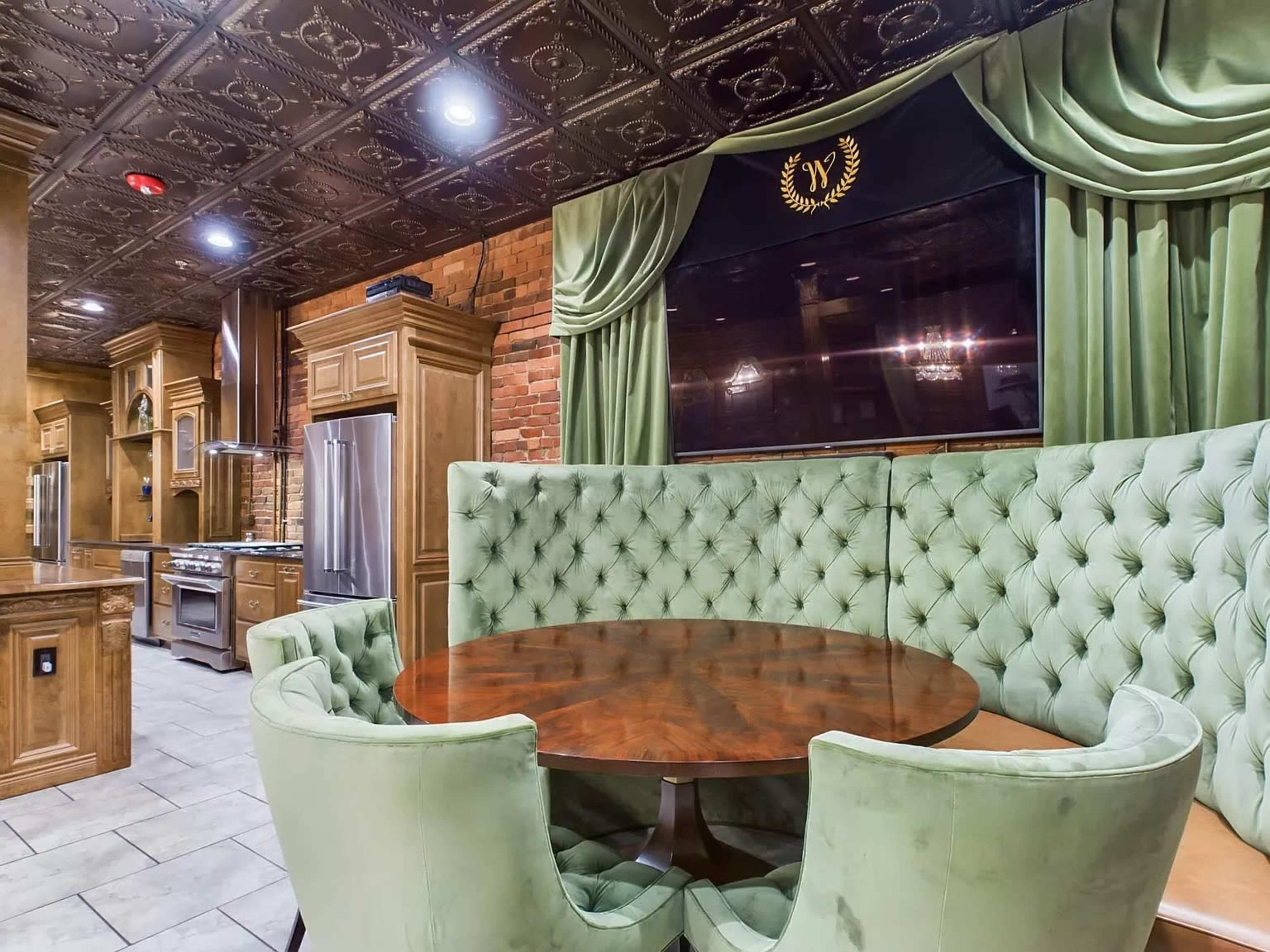 The image shows a kitchen area featuring a round wooden table surrounded by tufted green chairs, with a brick wall and decorative ceiling above.