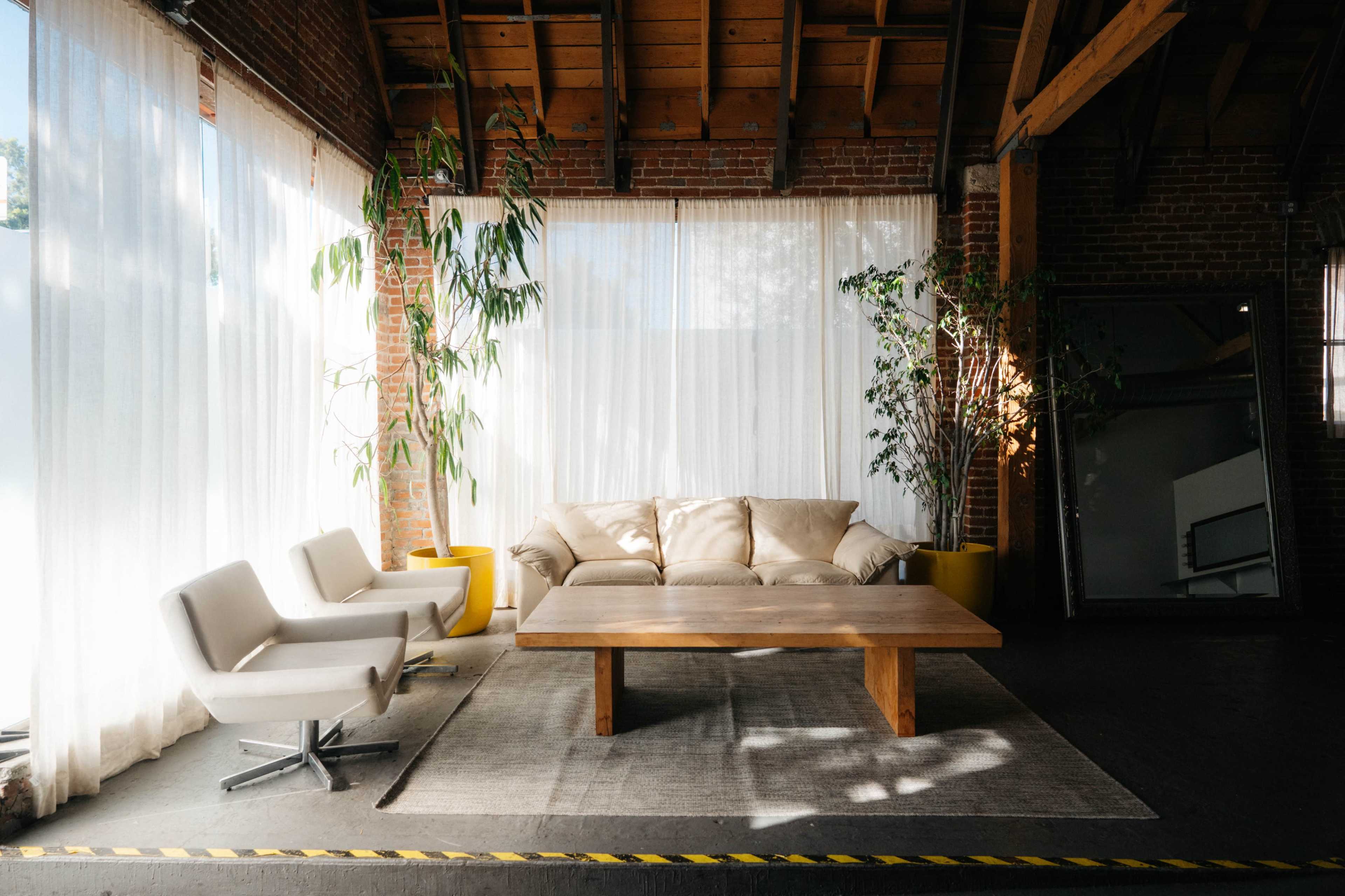 A minimalist living space features a beige sofa, a wooden coffee table, and two white swivel chairs, surrounded by large windows with sheer curtains and potted plants.