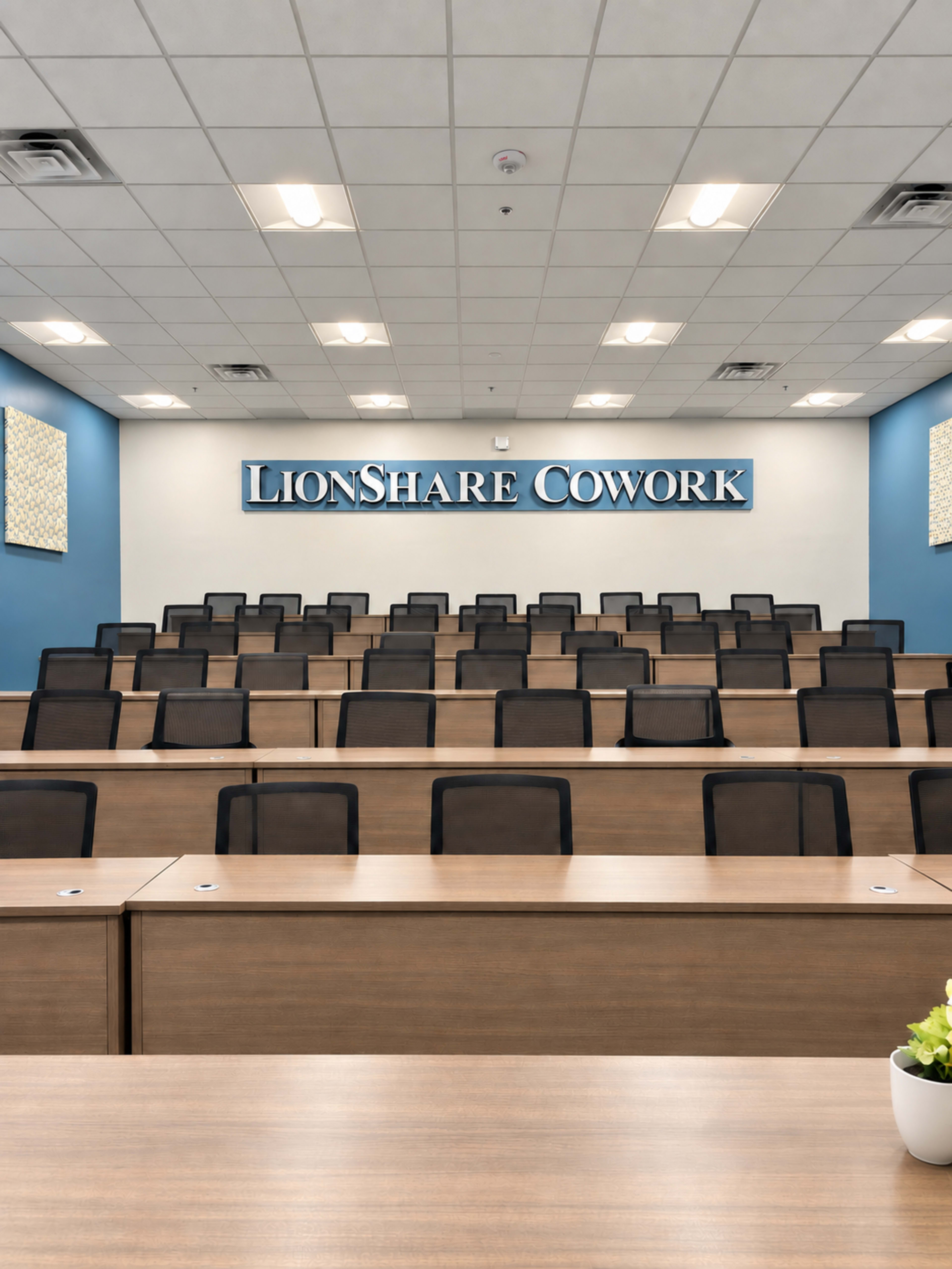 The image shows a spacious conference room with tiered seating, a large sign displaying "LionShare Cowork," and a well-lit ceiling.