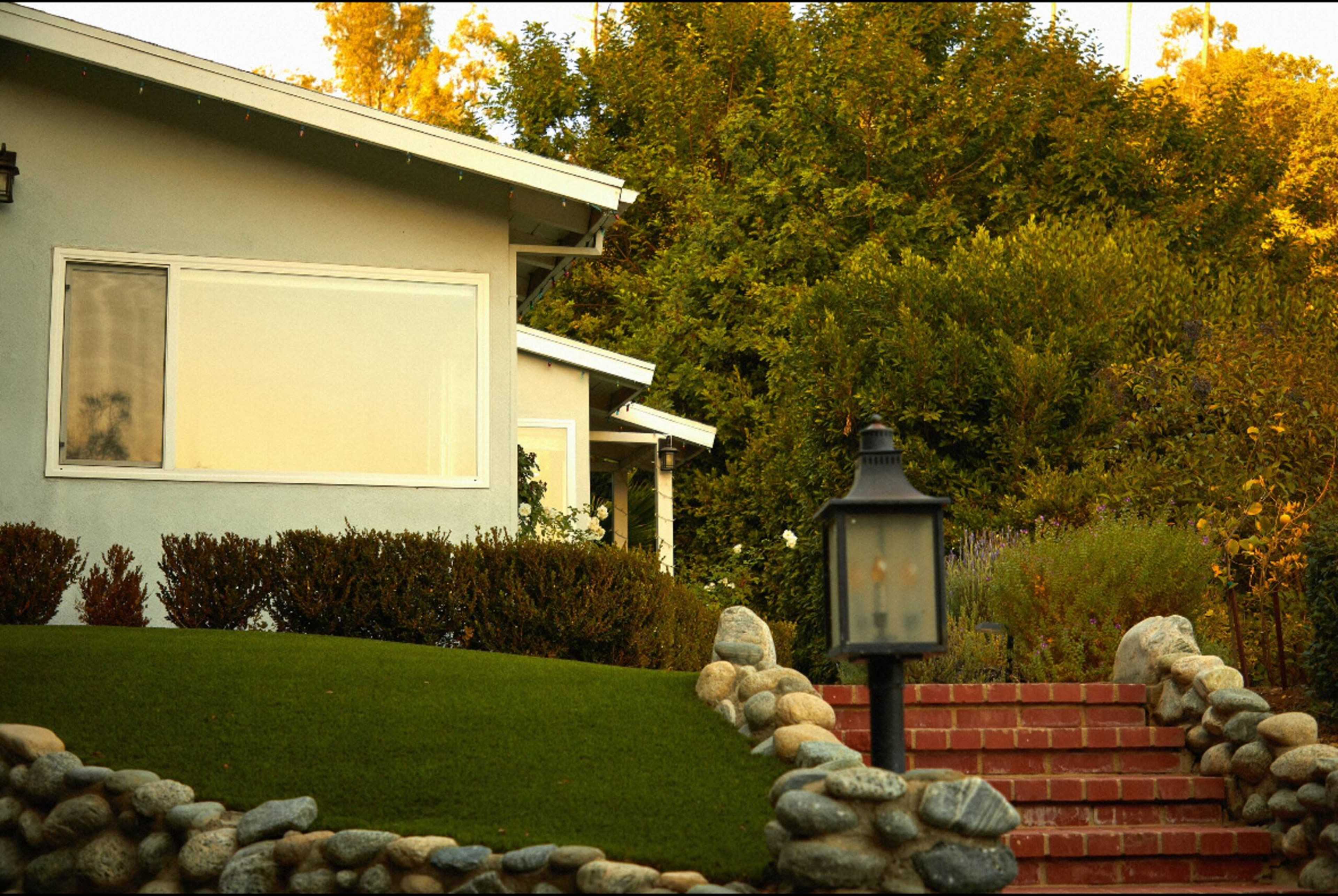 A light-colored house with a large window is situated beside a neatly landscaped lawn and a stone pathway leading to a set of brick steps.