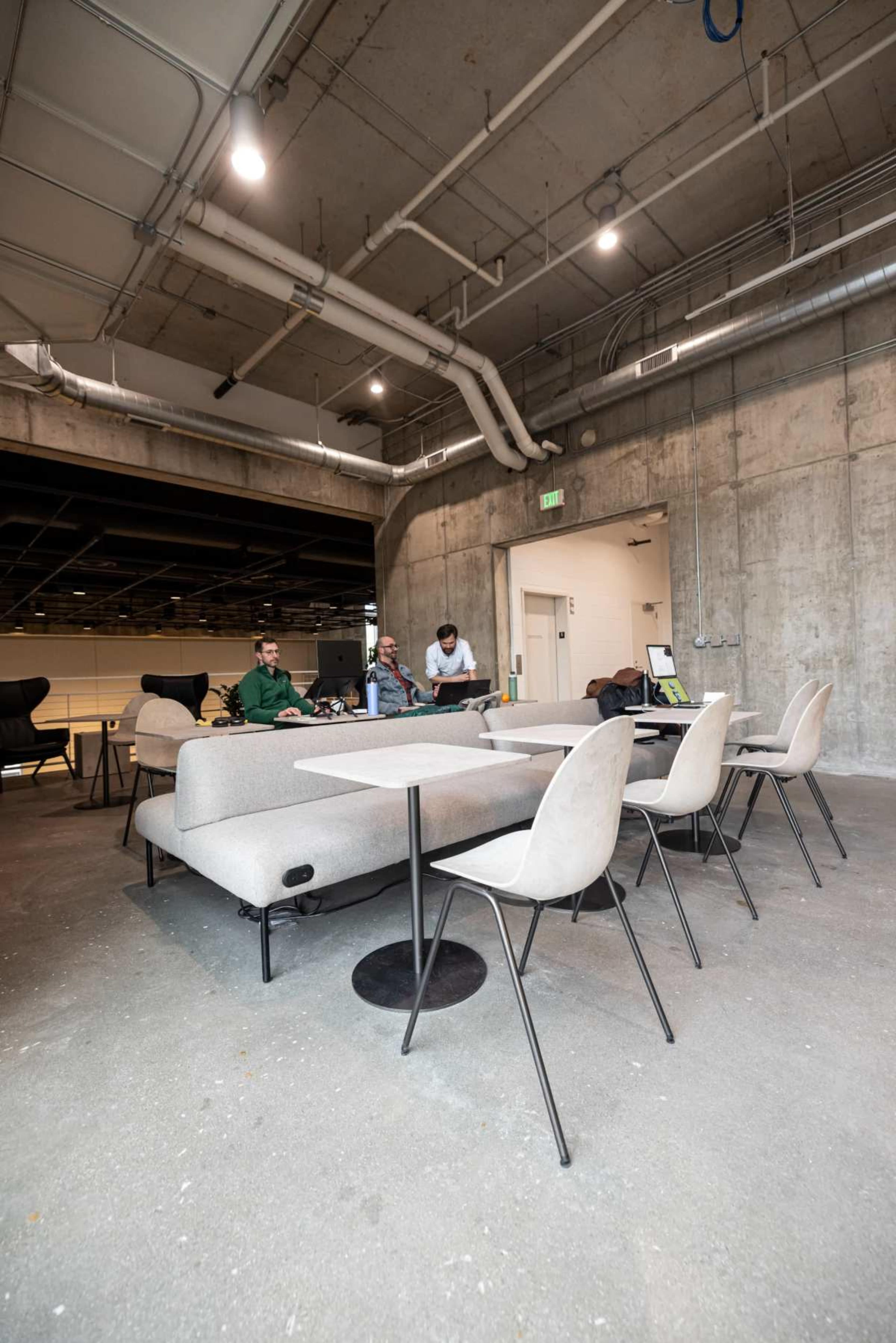 The image shows a modern, open workspace with gray sofas and chairs arranged around a low table, featuring exposed concrete walls and overhead piping.