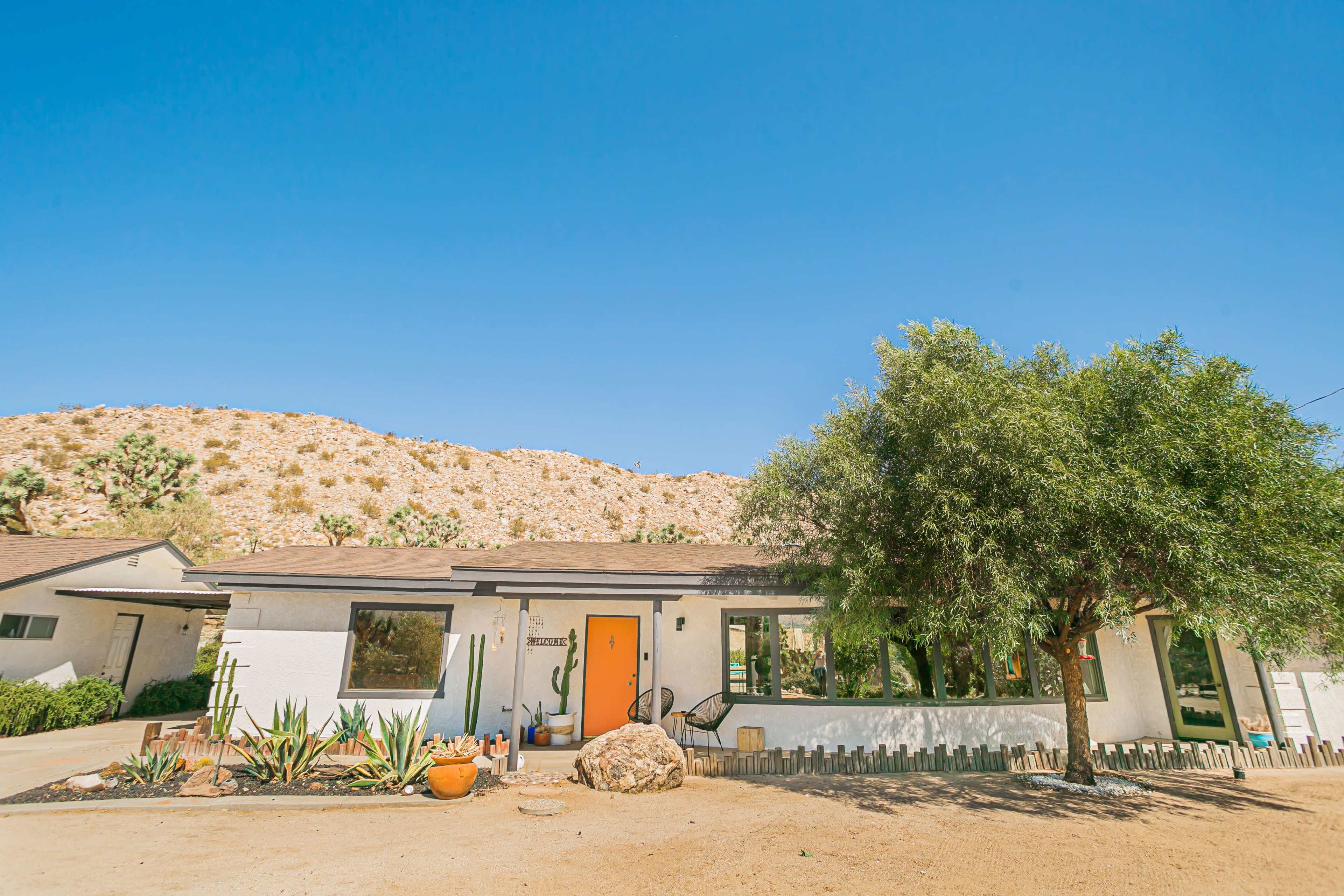A single-story white house with an orange door is situated in a desert landscape, framed by a rocky hillside and greenery.