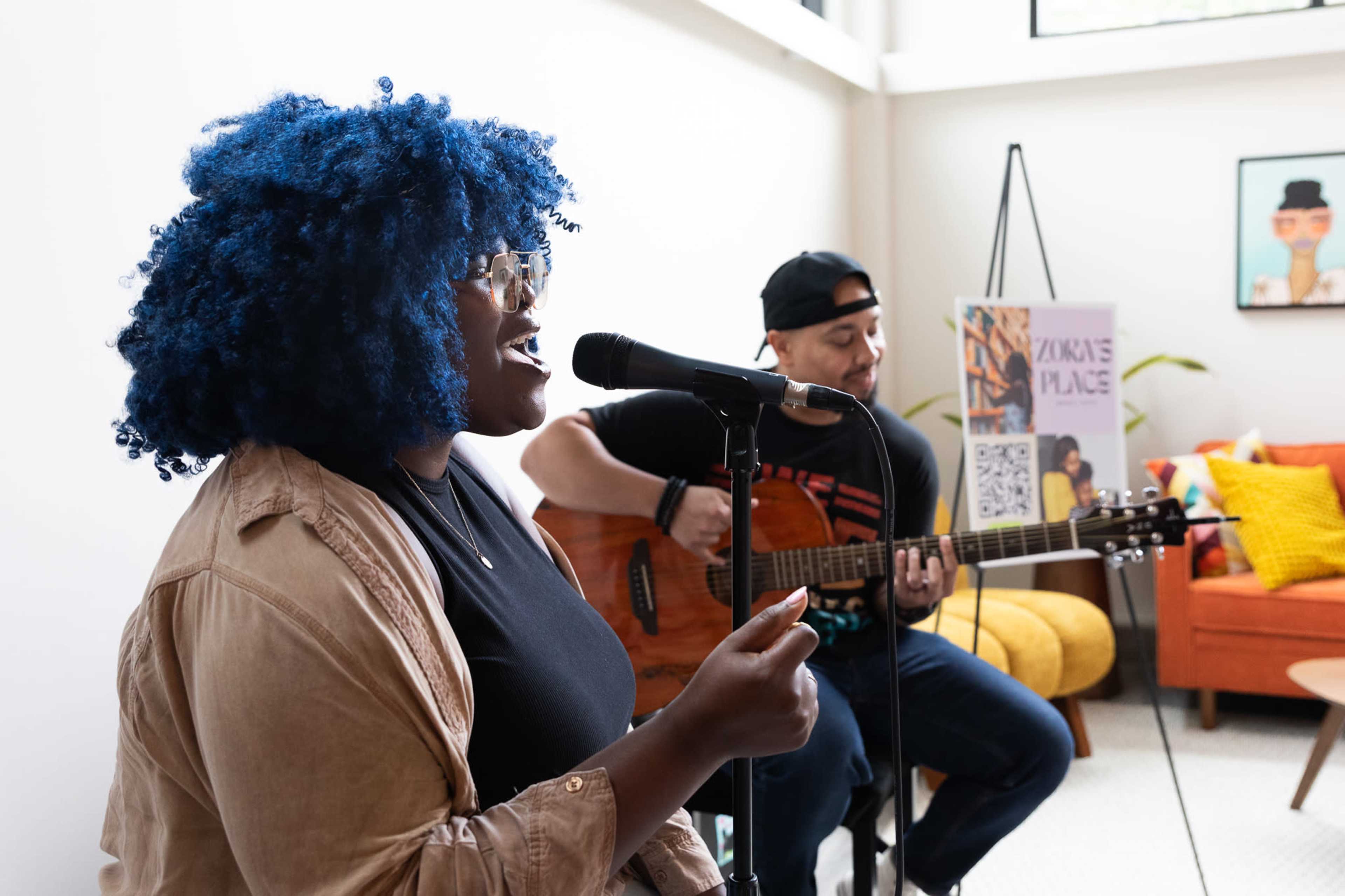 A woman with blue curly hair sings into a microphone while a man plays guitar beside her in a well-lit indoor space.