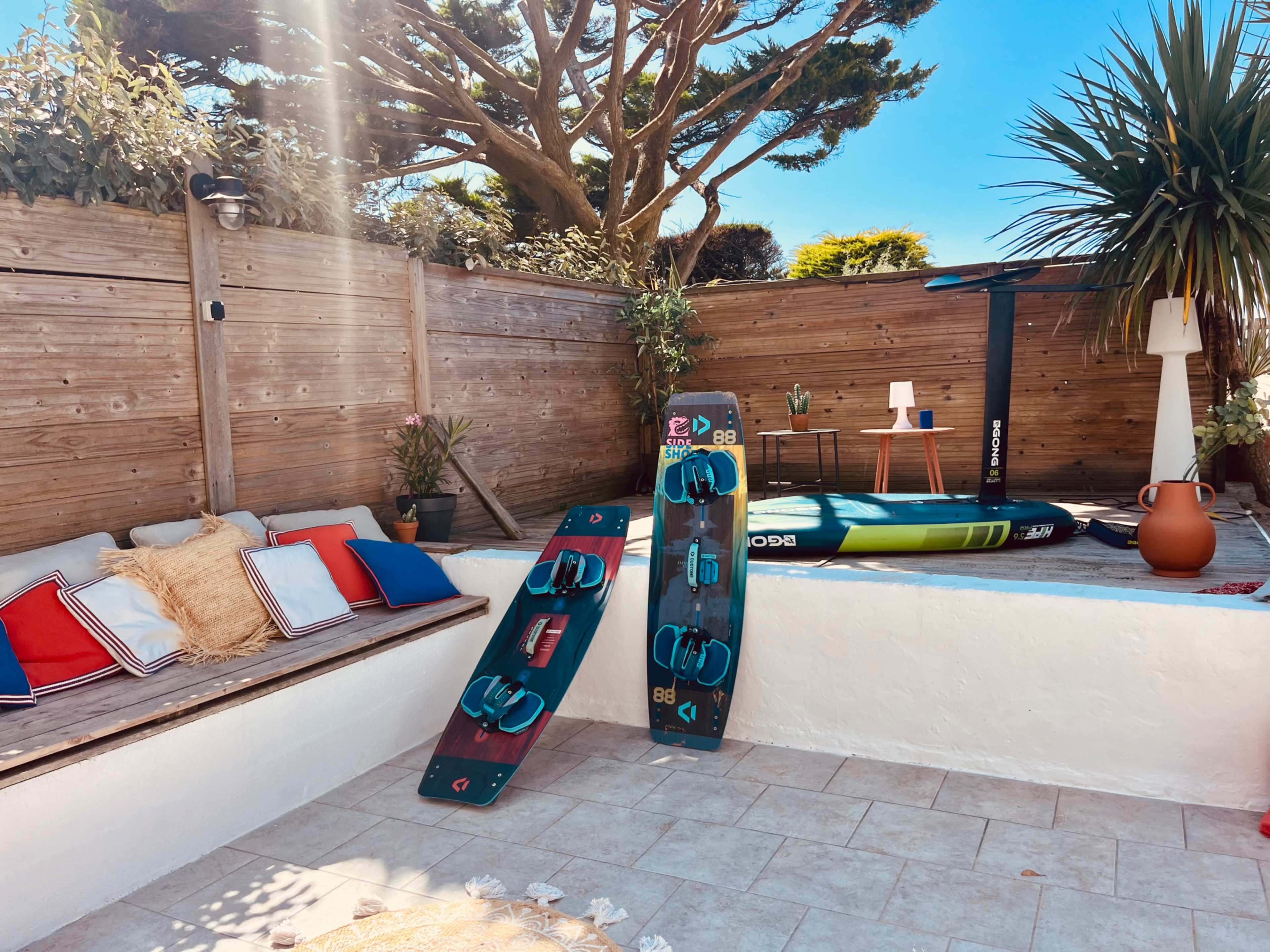 The image shows a sunny outdoor space with two kiteboards propped against a wall, a paddleboard resting in the background, and a small seating area decorated with cushions and a palm tree.