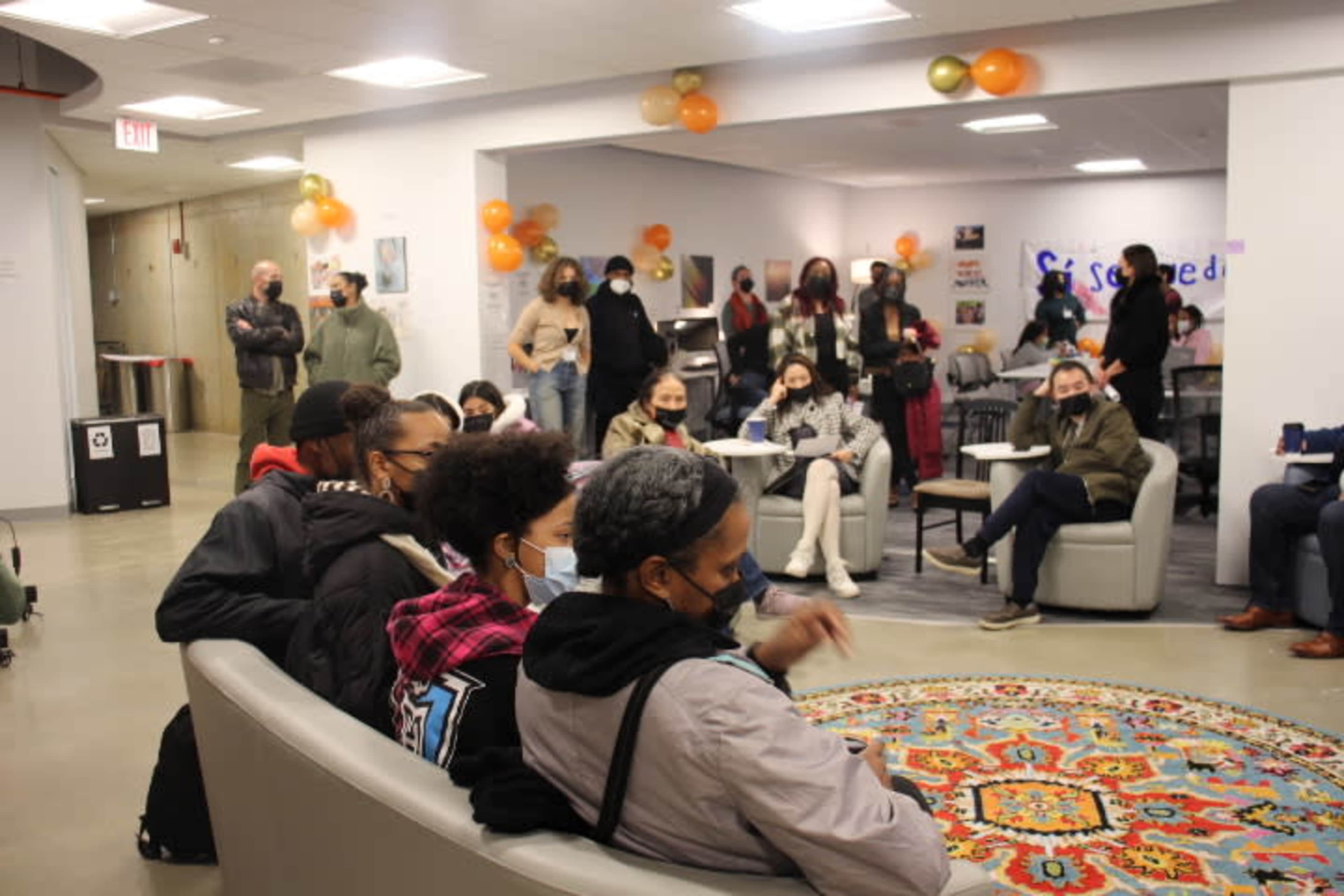 A group of people wearing masks are seated in a lounge area with chairs and a large, colorful rug, while others stand and engage in conversation.