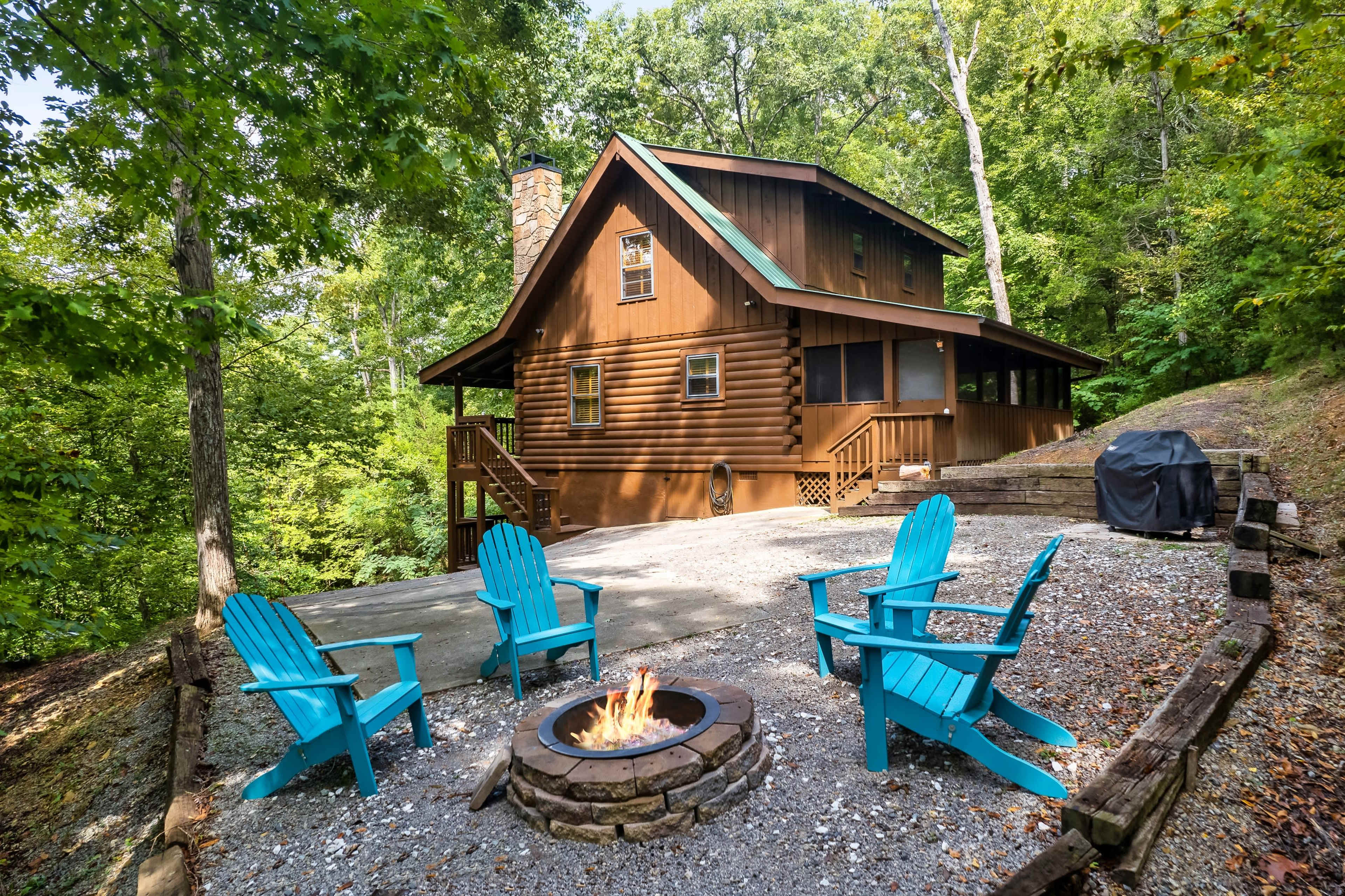 A two-story log cabin sits nestled among trees, with a fire pit and blue Adirondack chairs arranged around it in a gravel area.