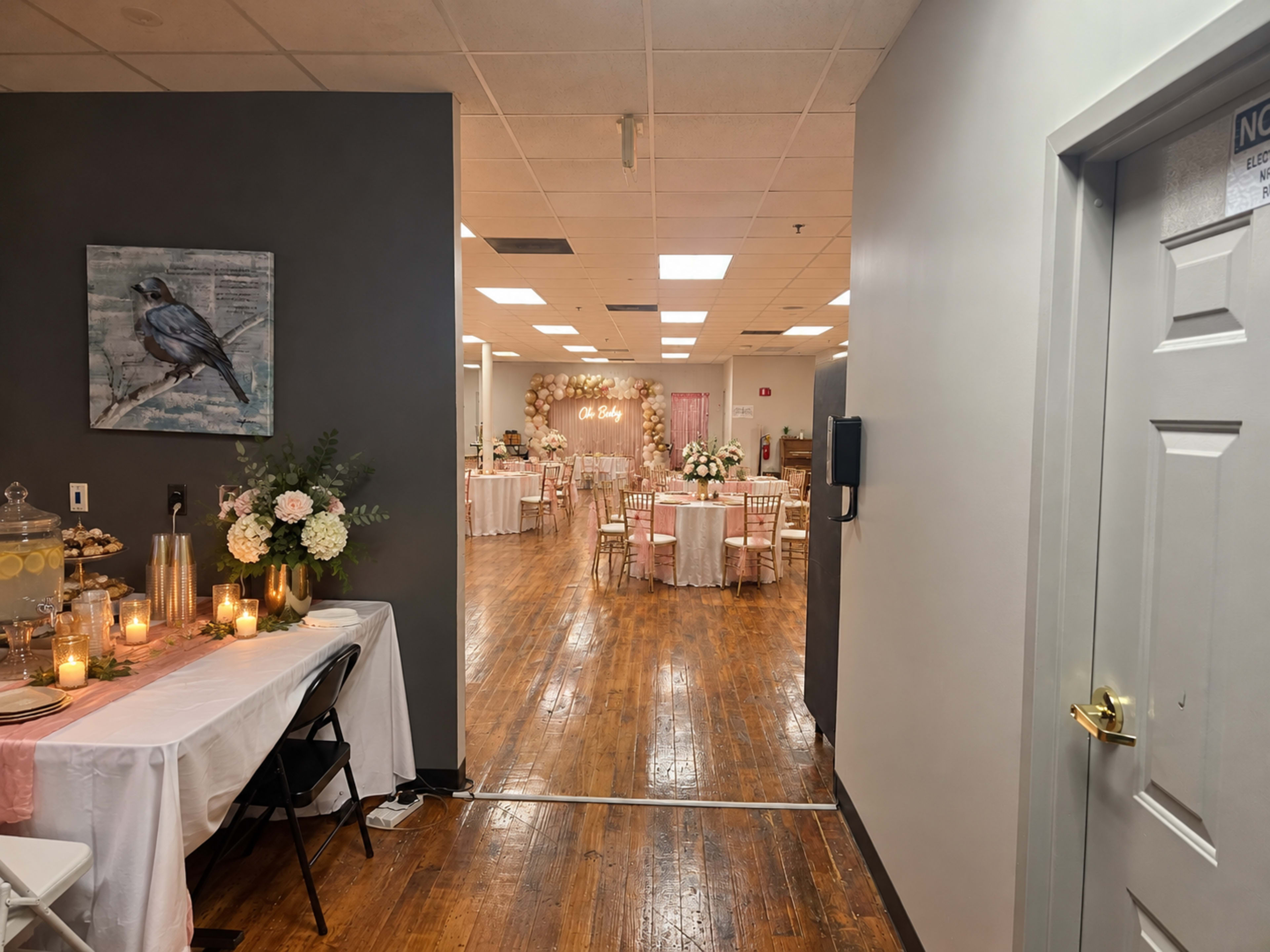 The image shows a spacious event venue with tables set for a celebration, decorative floral arrangements, and a backdrop for photographs.
