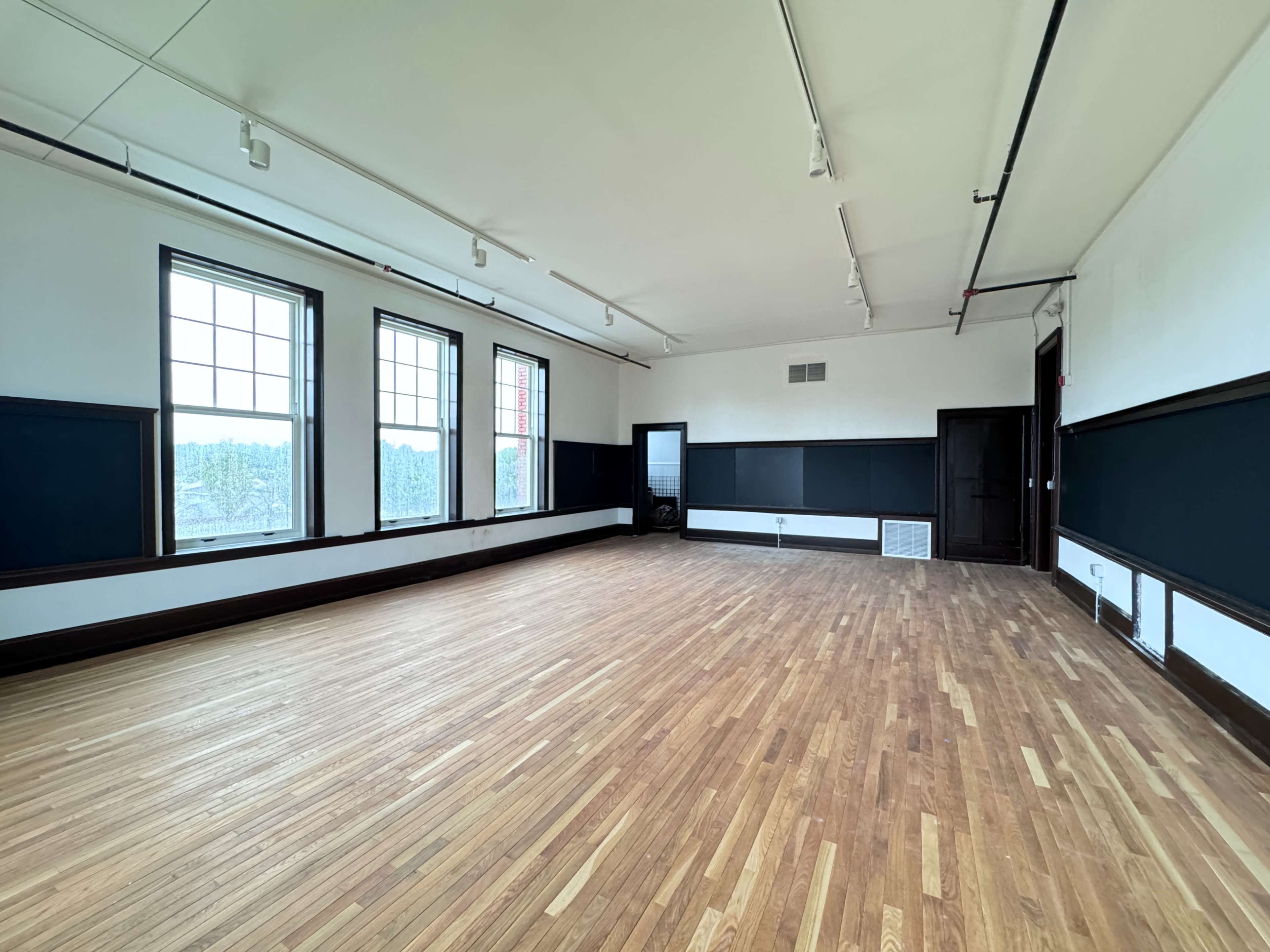 The image shows a spacious, empty room with wooden flooring, large windows, and black-painted walls.