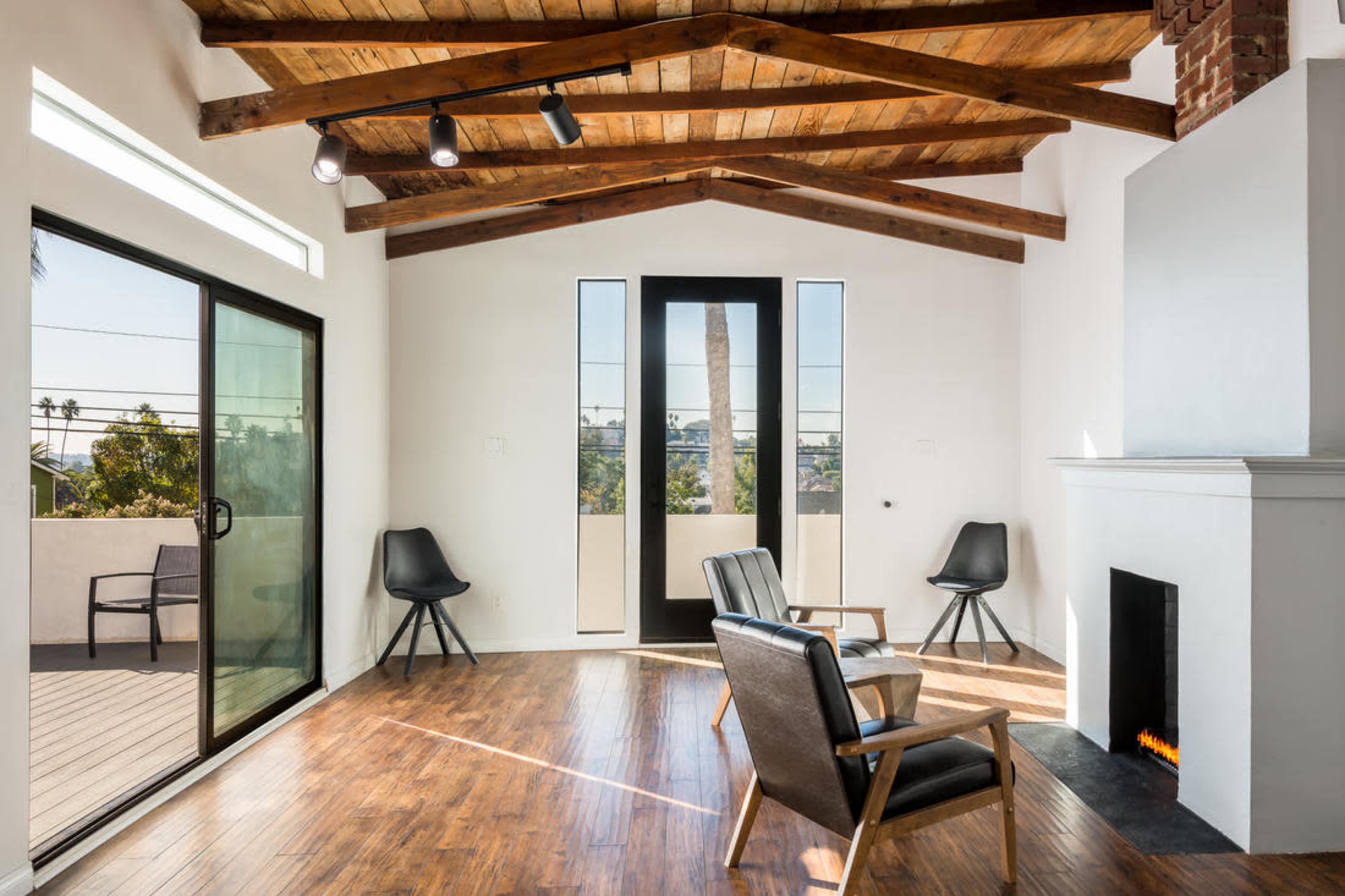A modern interior living space with wooden beams, a brick fireplace, and large windows facing a patio.