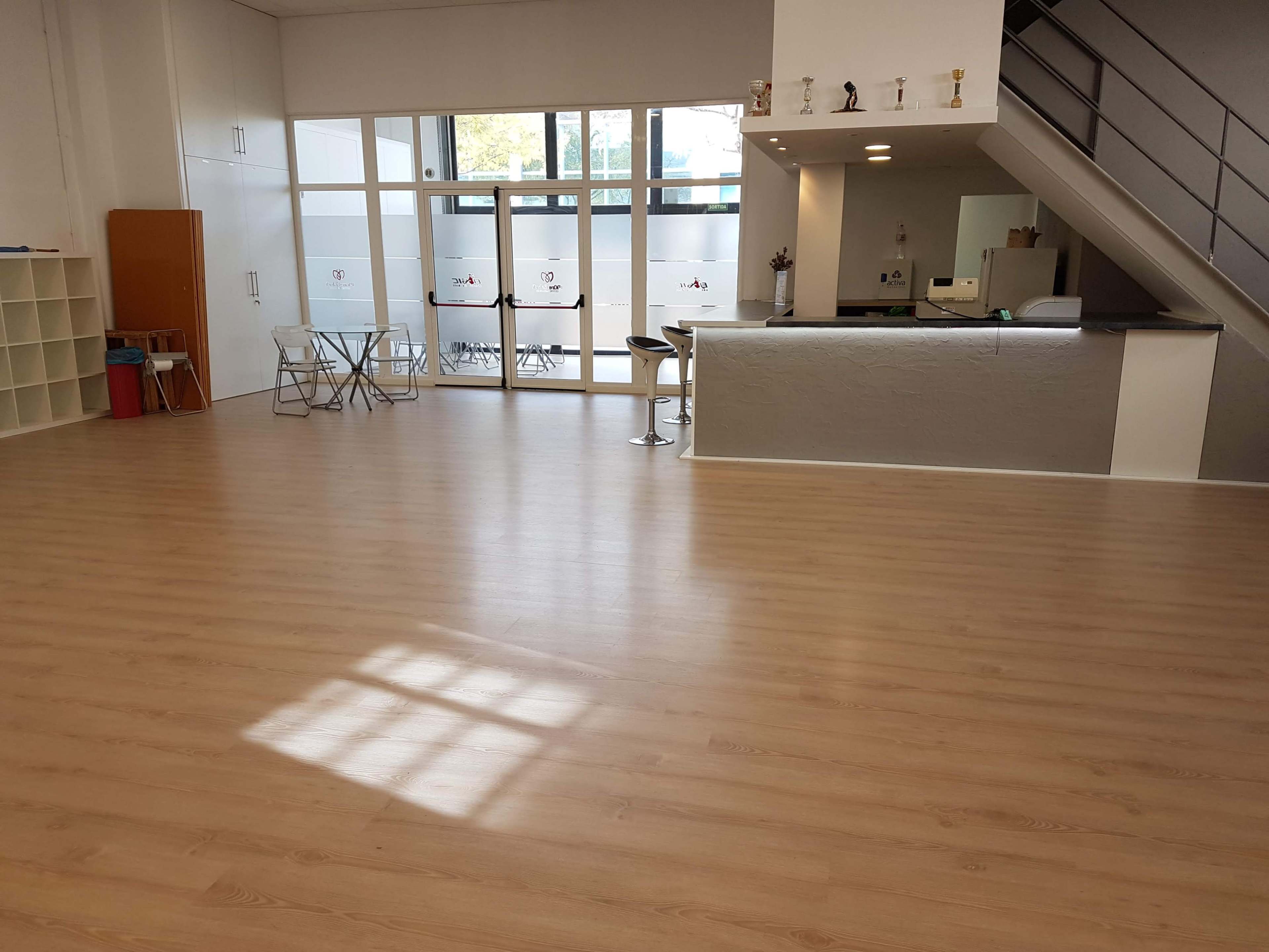 A spacious indoor area with light wood flooring, two chairs and a table, and a bar-like counter visible behind a glass door.