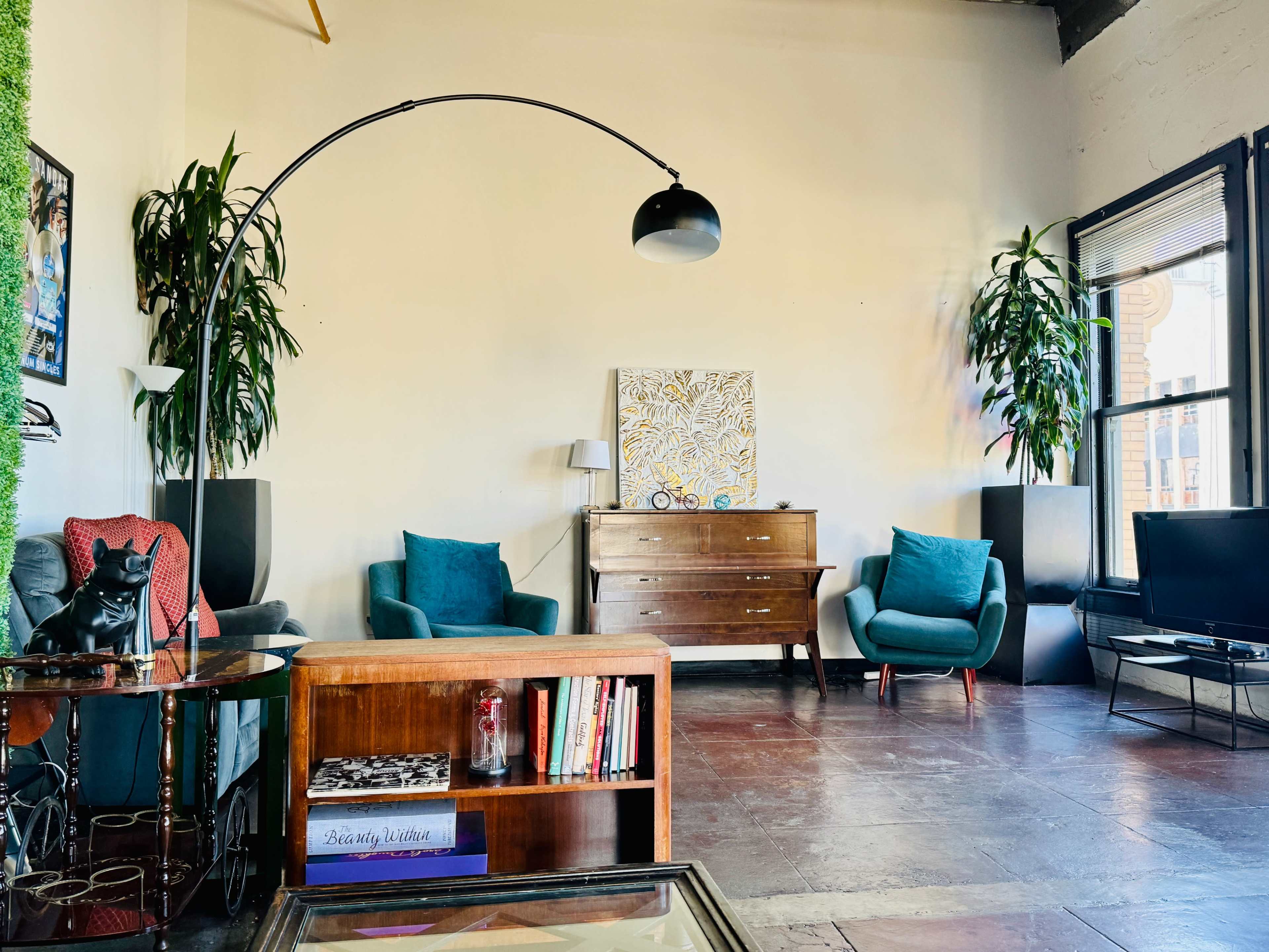A bright living room featuring two teal armchairs, a wooden sideboard, a black television, and indoor plants.