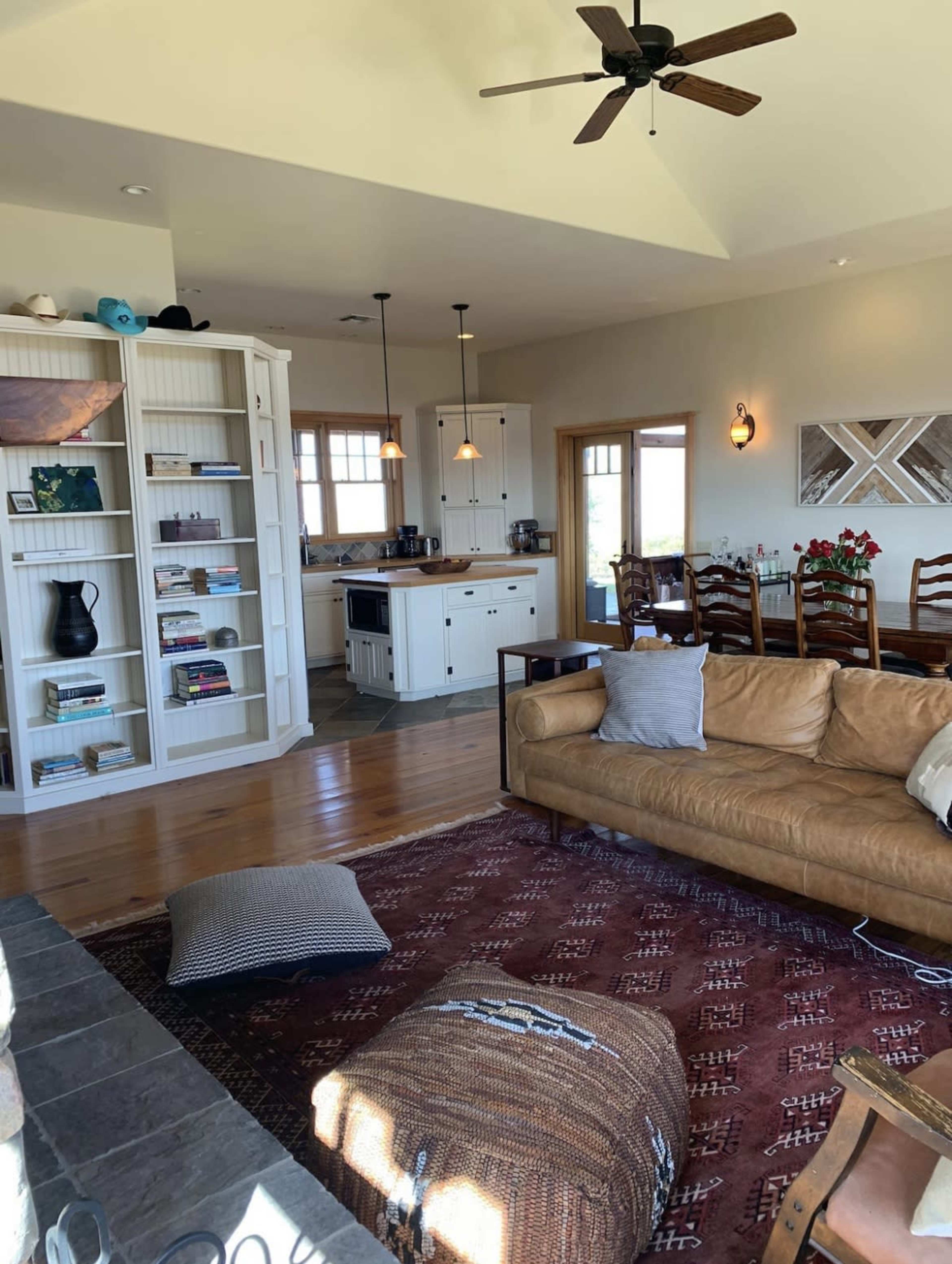 A spacious living area featuring a leather sofa, a large area rug, a wooden dining table, and a kitchen in the background with white cabinetry.