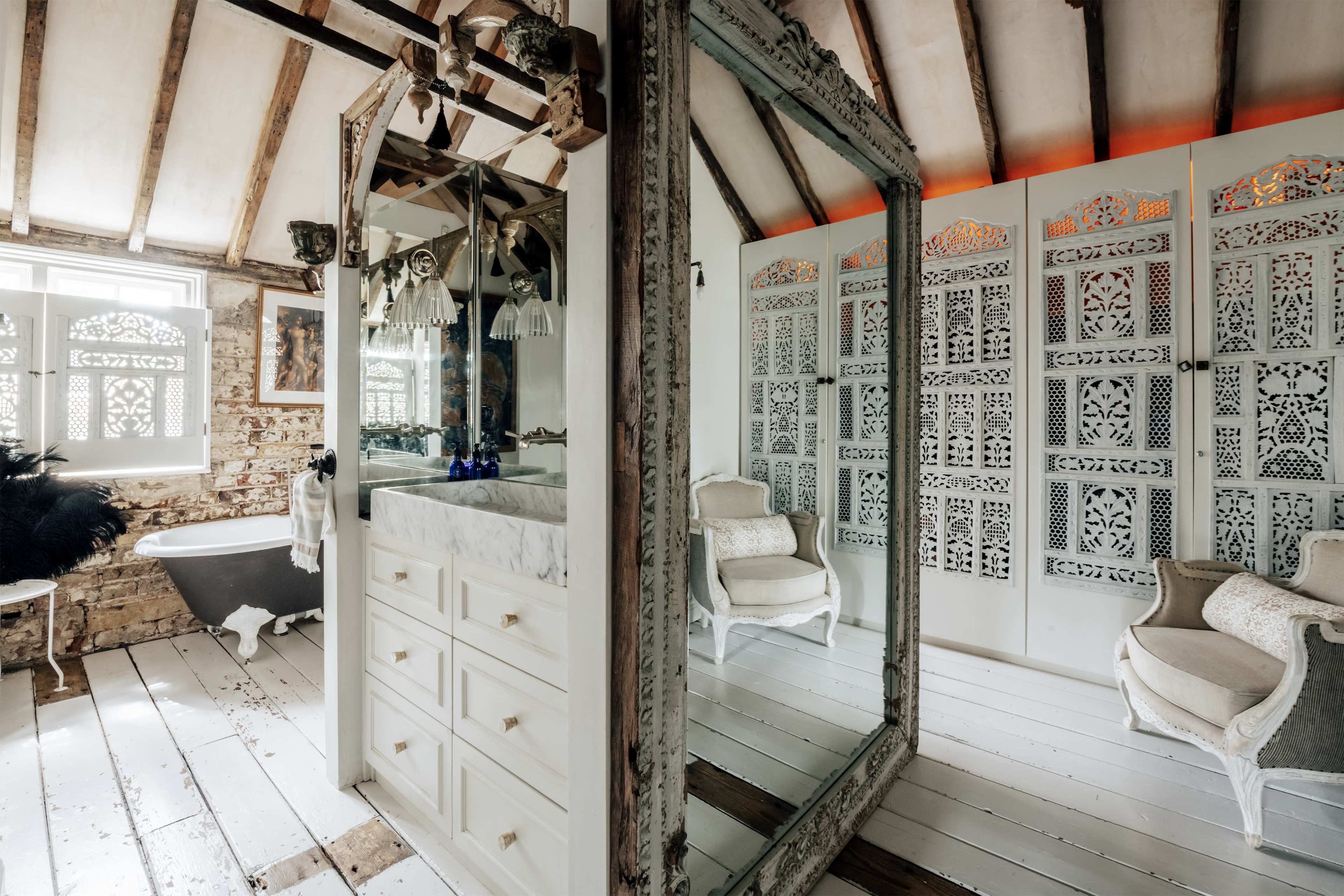 The image shows a rustic bathroom featuring a freestanding bathtub, a modern sink with cabinetry, and a large ornate mirror, all set against exposed wooden beams and intricate decorative screens.