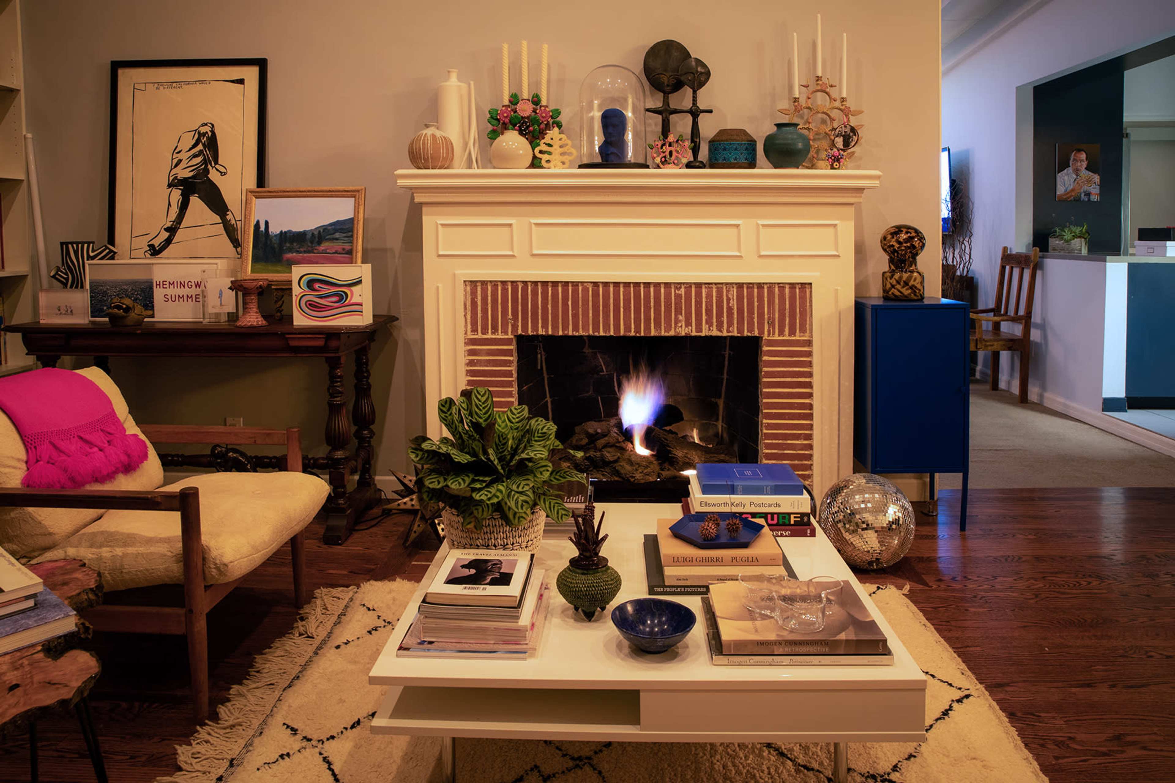 A cozy living room features a fireplace with an open flame, surrounded by decorative items, books, and a potted plant on a coffee table.
