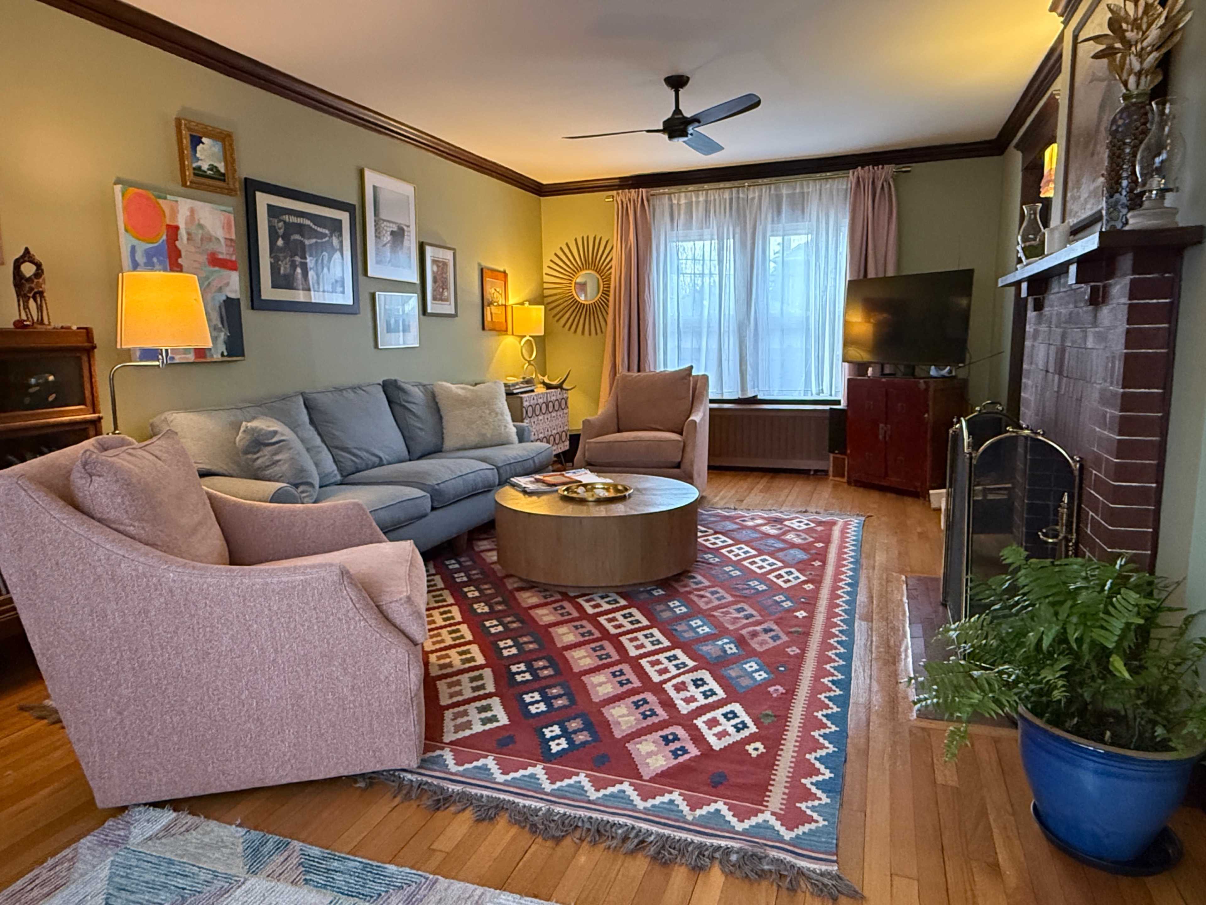 The living room features a grey sectional sofa, two pink armchairs, a round wooden coffee table on a patterned rug, and a window with sheer curtains.