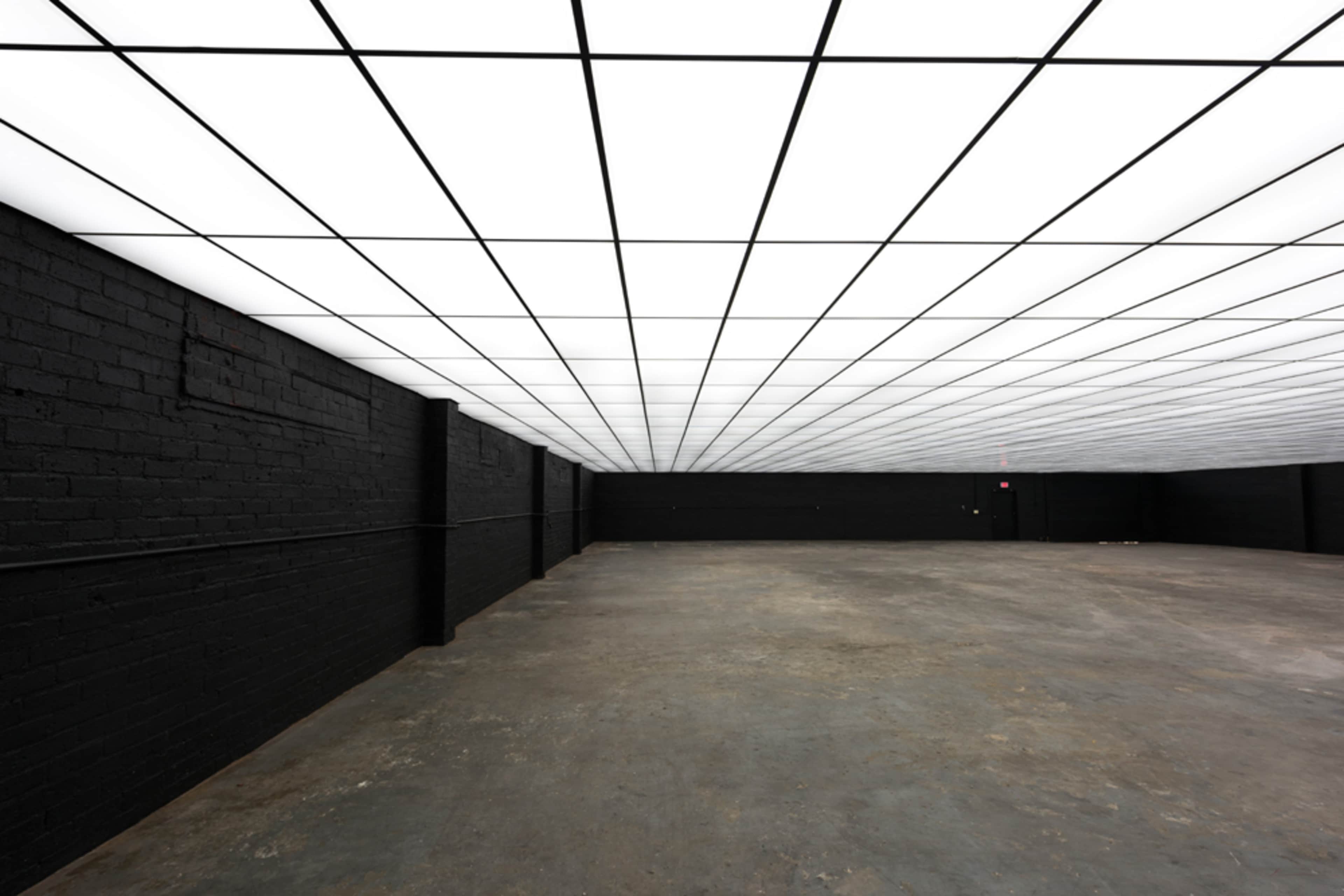 A large, empty room with black walls and a grid-patterned ceiling composed of bright, fluorescent lights.