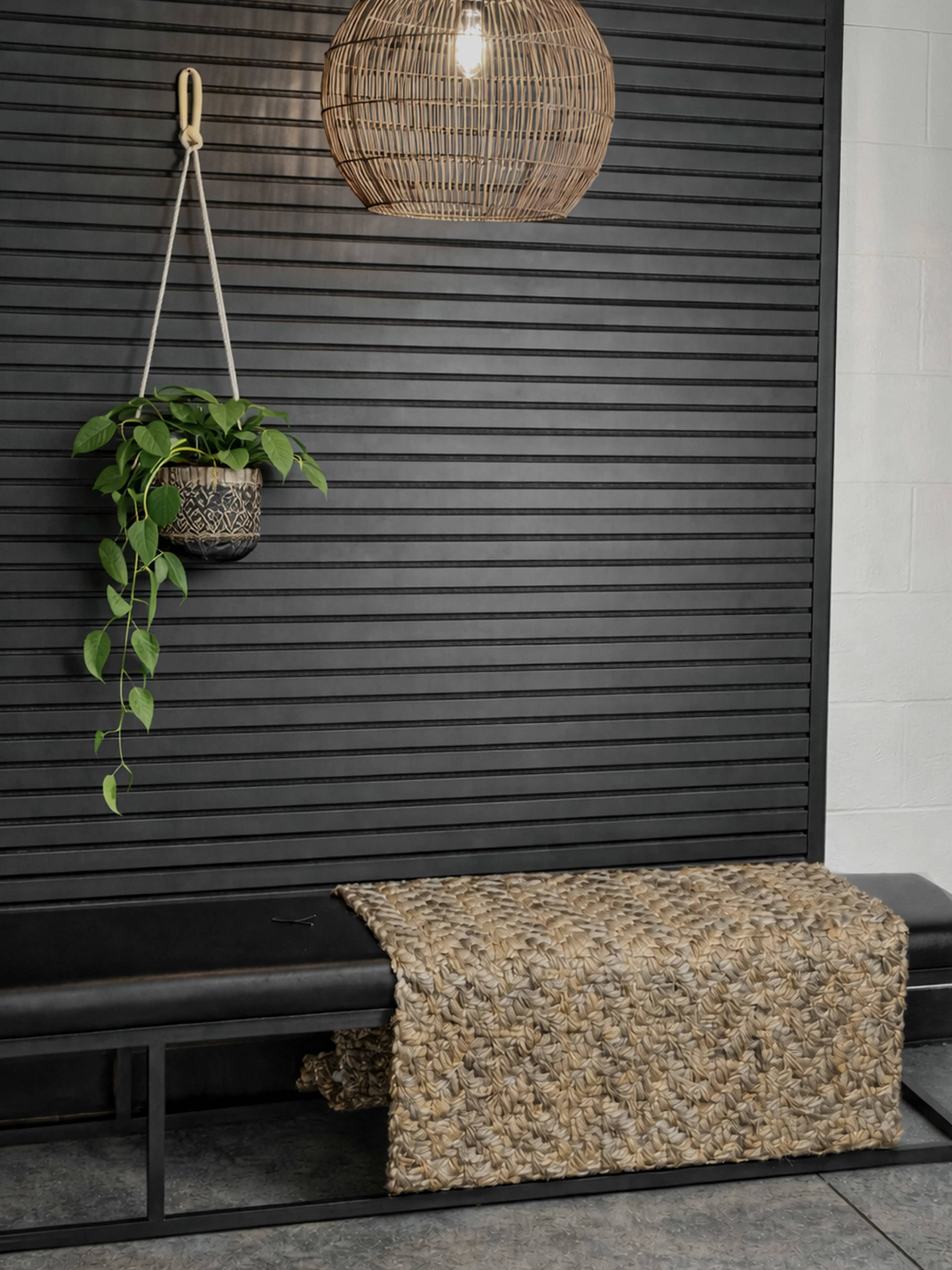 The image features a woven bench adorned with a textured mat, positioned against a black slatted wall with a hanging rattan light fixture and a planter.