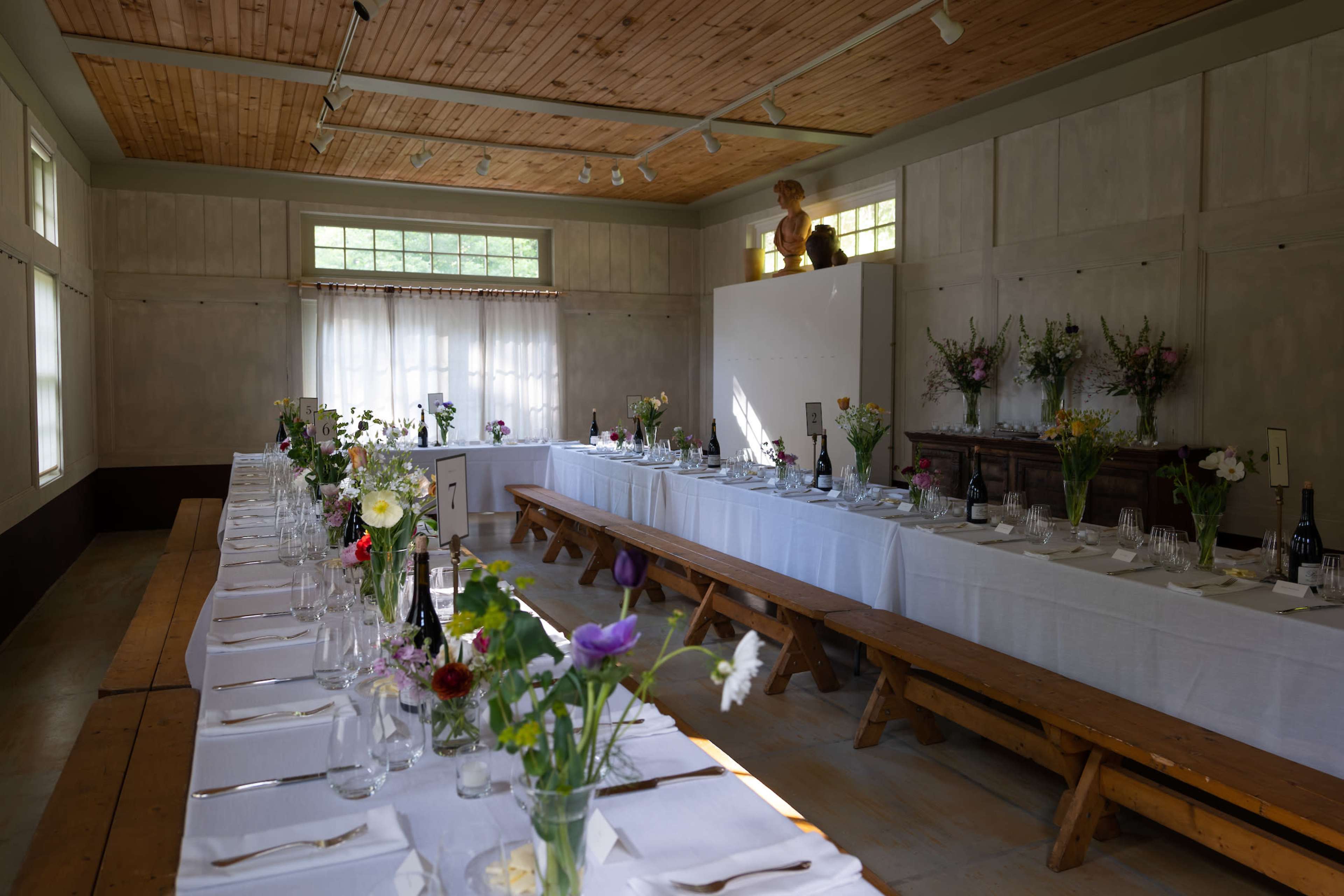 A long dining table set with white tablecloths and glassware, decorated with flowers, is positioned in a well-lit room with wooden ceilings and benches.