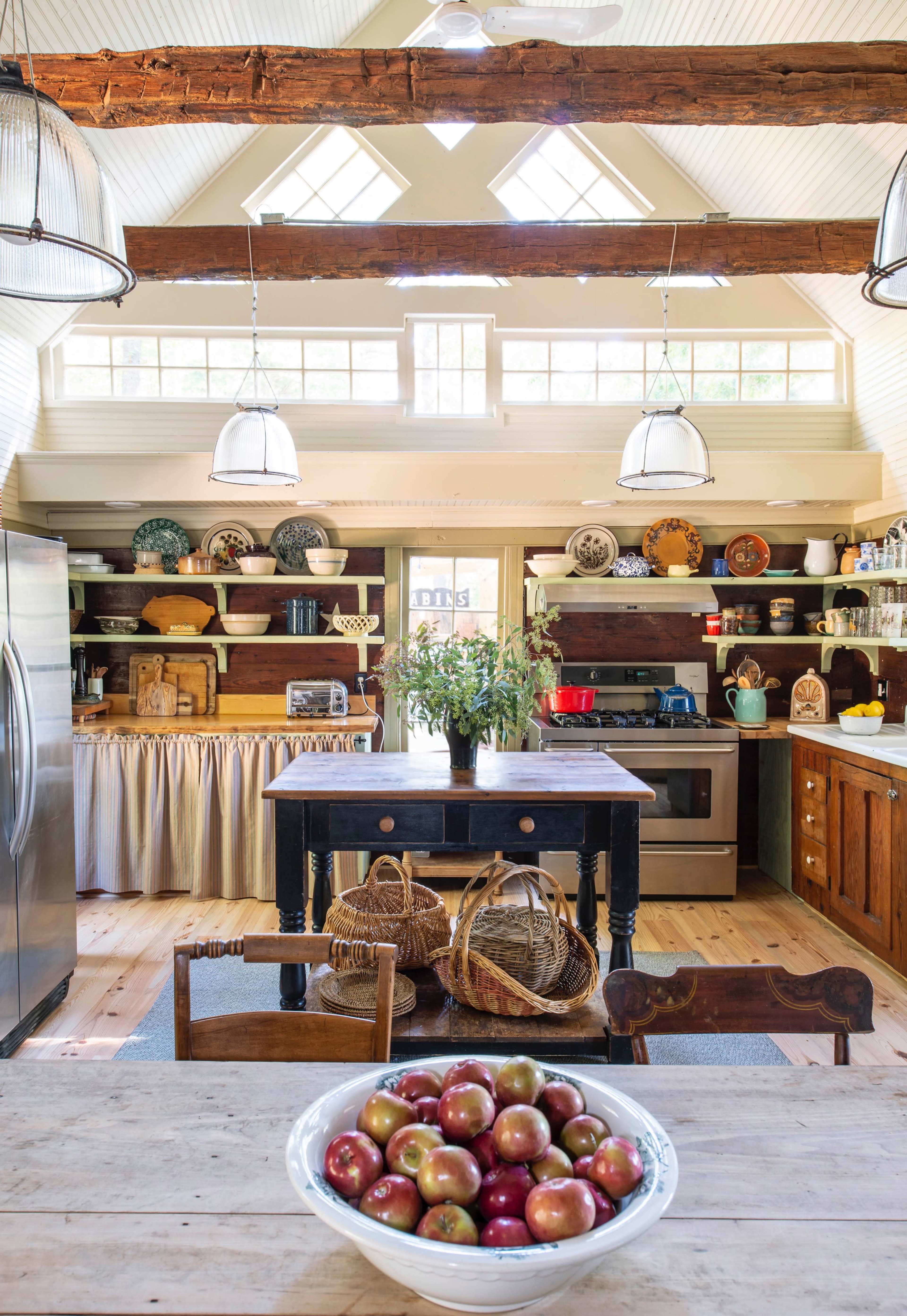 A spacious kitchen with wooden beams features a central table with a bowl of apples and various shelves displaying dishes and cookware.