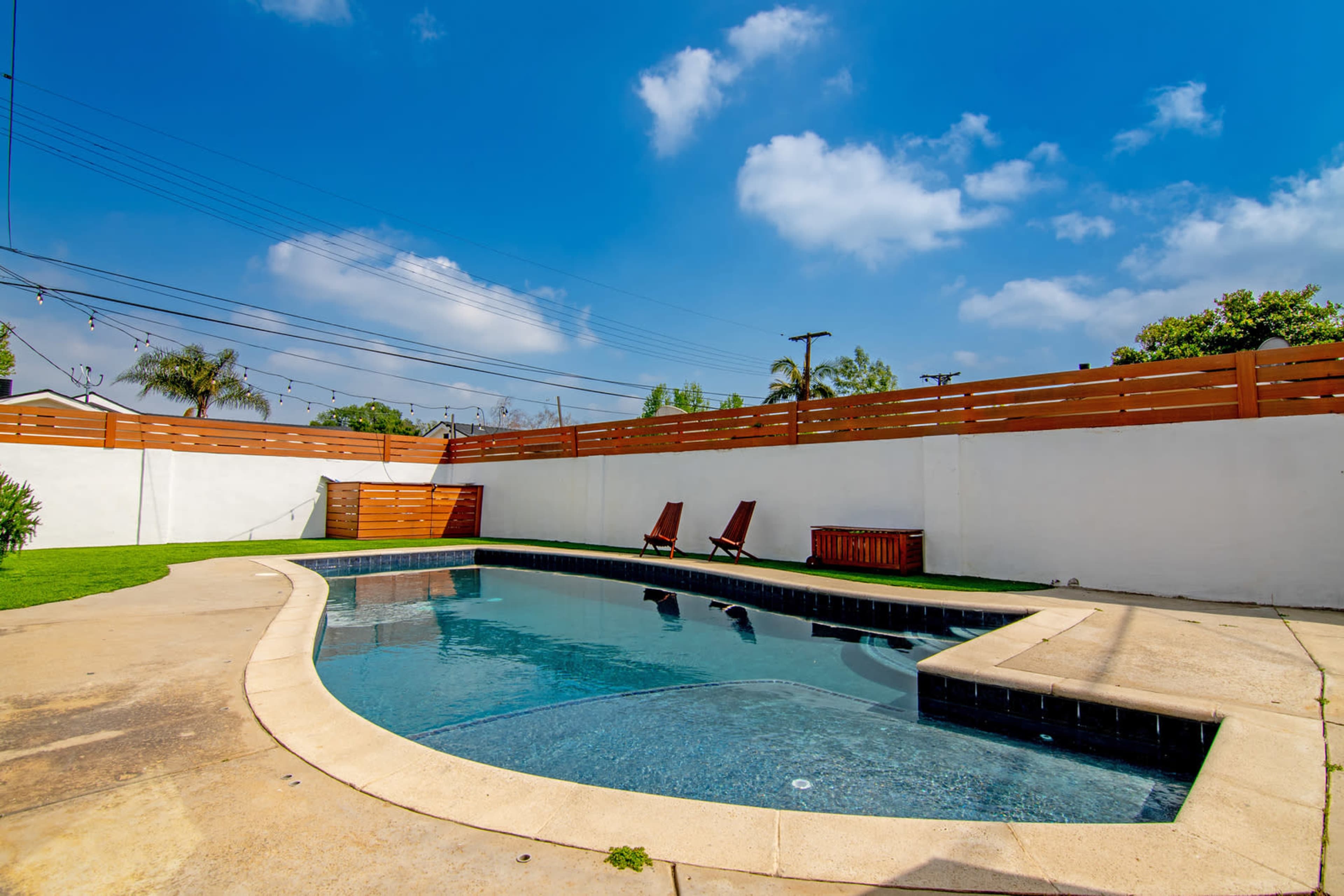 A swimming pool with curved edges is surrounded by a concrete deck and a green lawn, with two wooden lounge chairs on one side and a wooden storage bench nearby.