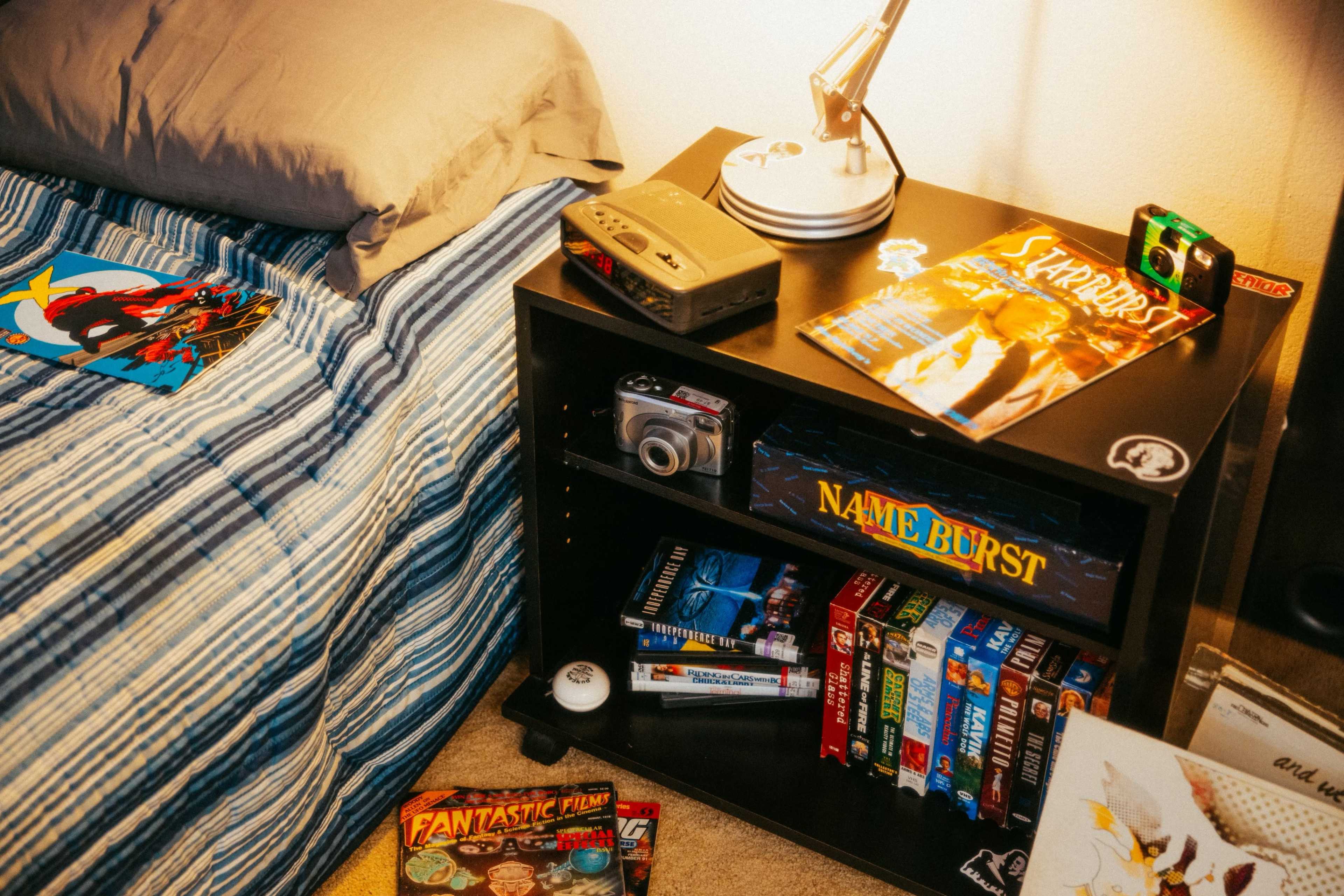 A bedside table holds various items, including magazines, a film camera, and VHS tapes, next to a neatly made bed with a striped comforter.