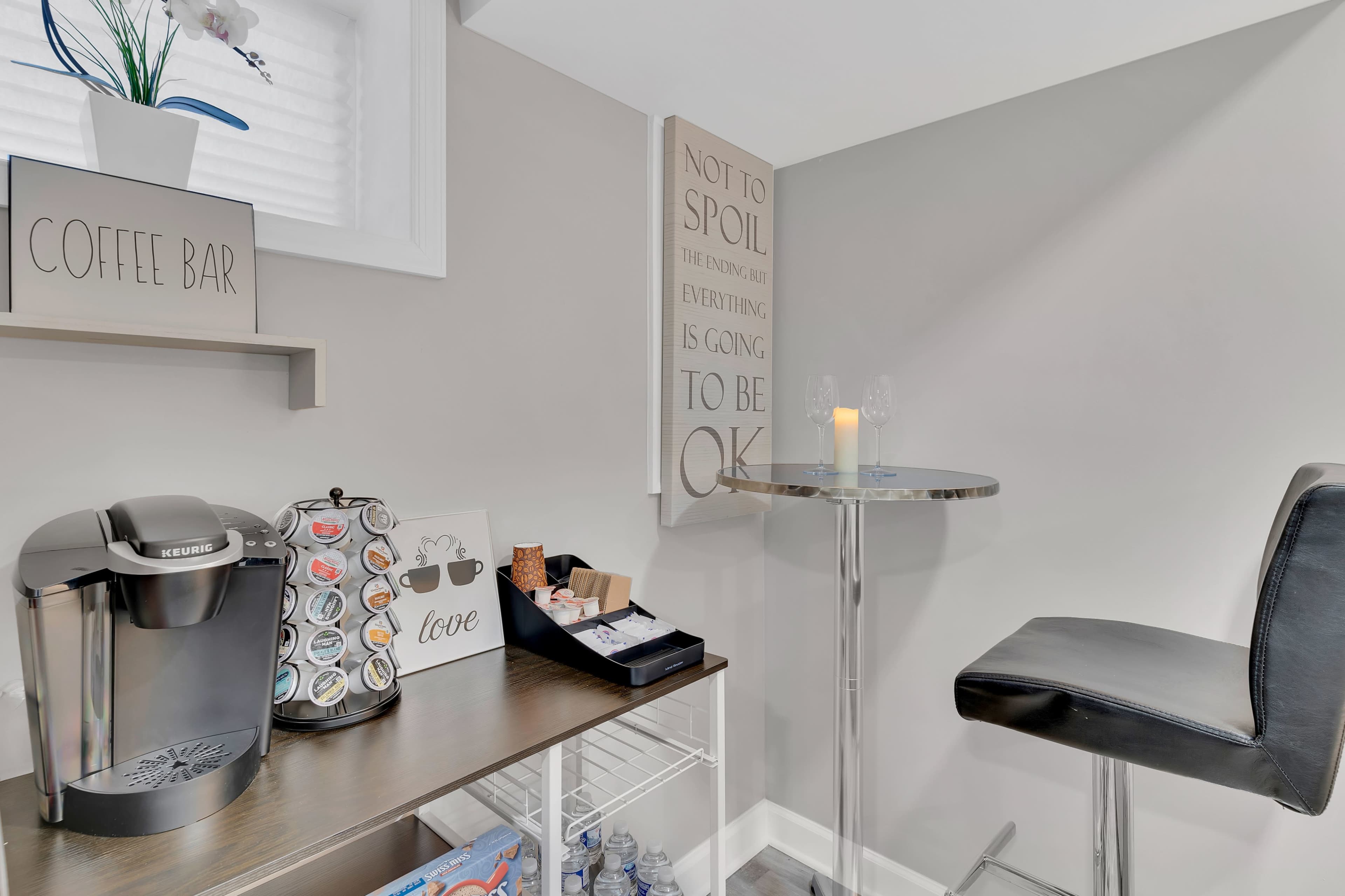 A corner of a room features a coffee bar station with a Keurig machine, a table, and some decorative items on the wall.