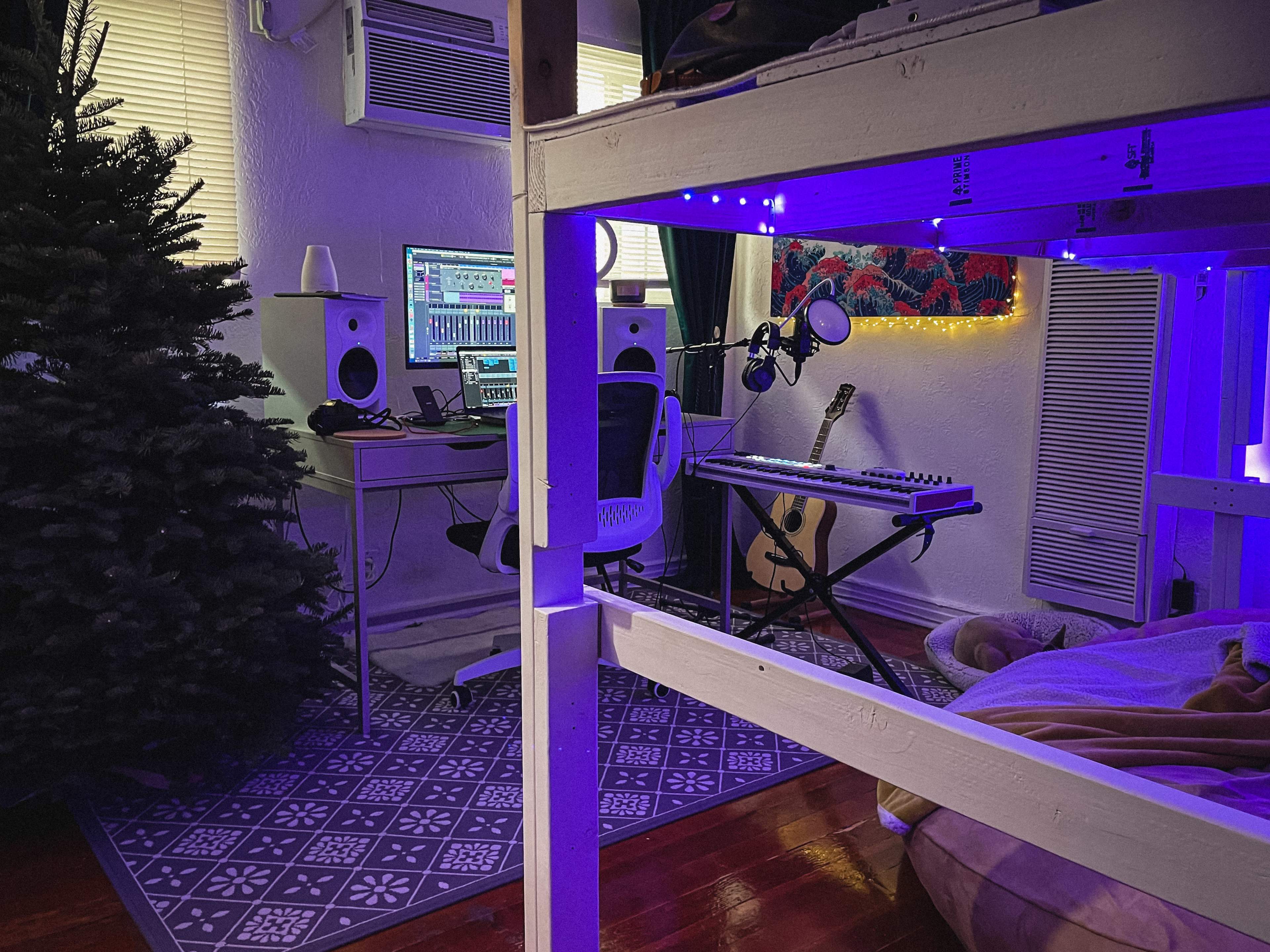 A dimly lit room features a loft bed, an air conditioning unit, a music workstation with speakers and a keyboard, and a Christmas tree in the corner.