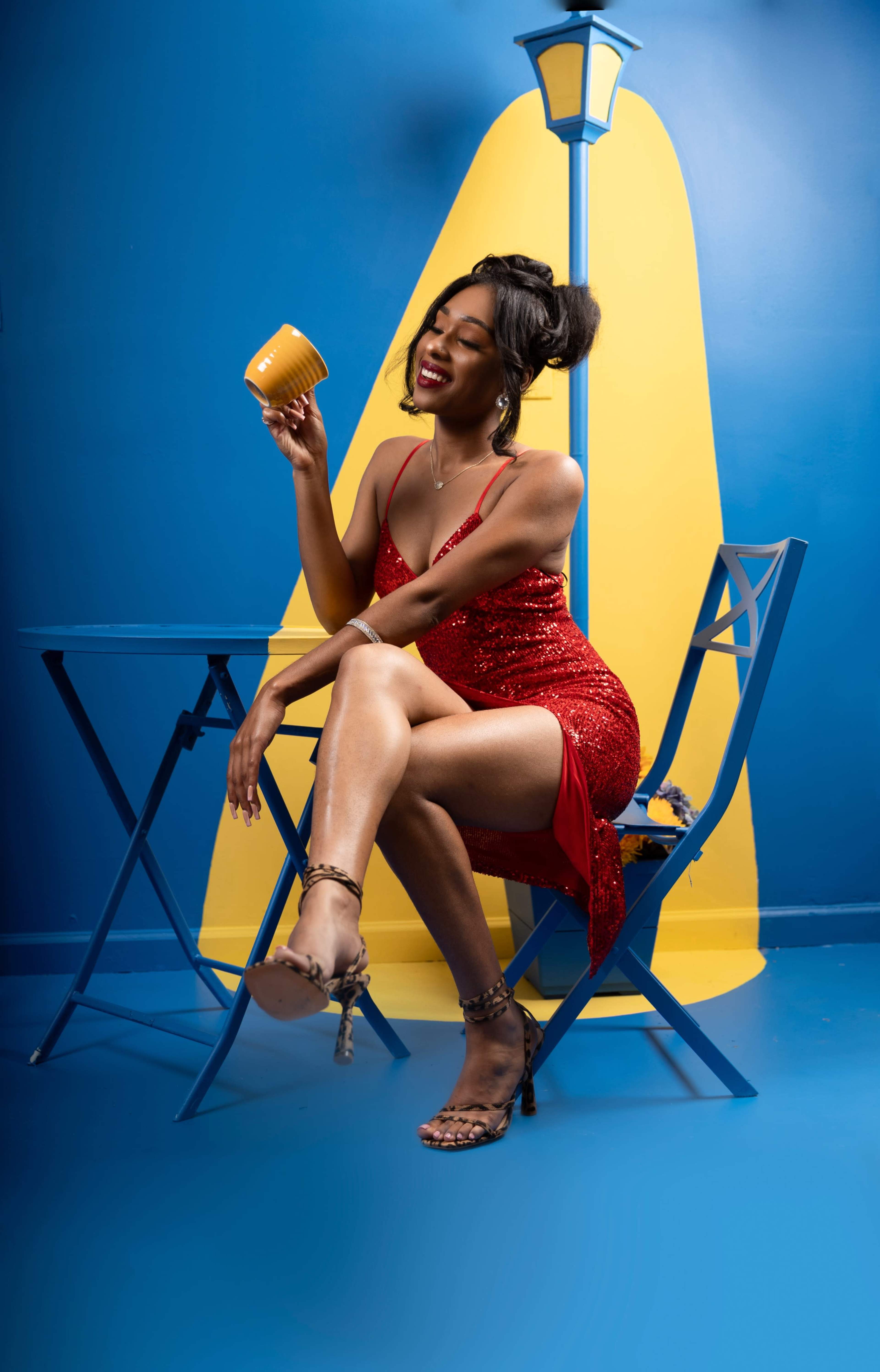 A woman in a red sequined dress sits on a blue chair at a small table, holding a yellow mug against a blue and yellow backdrop.