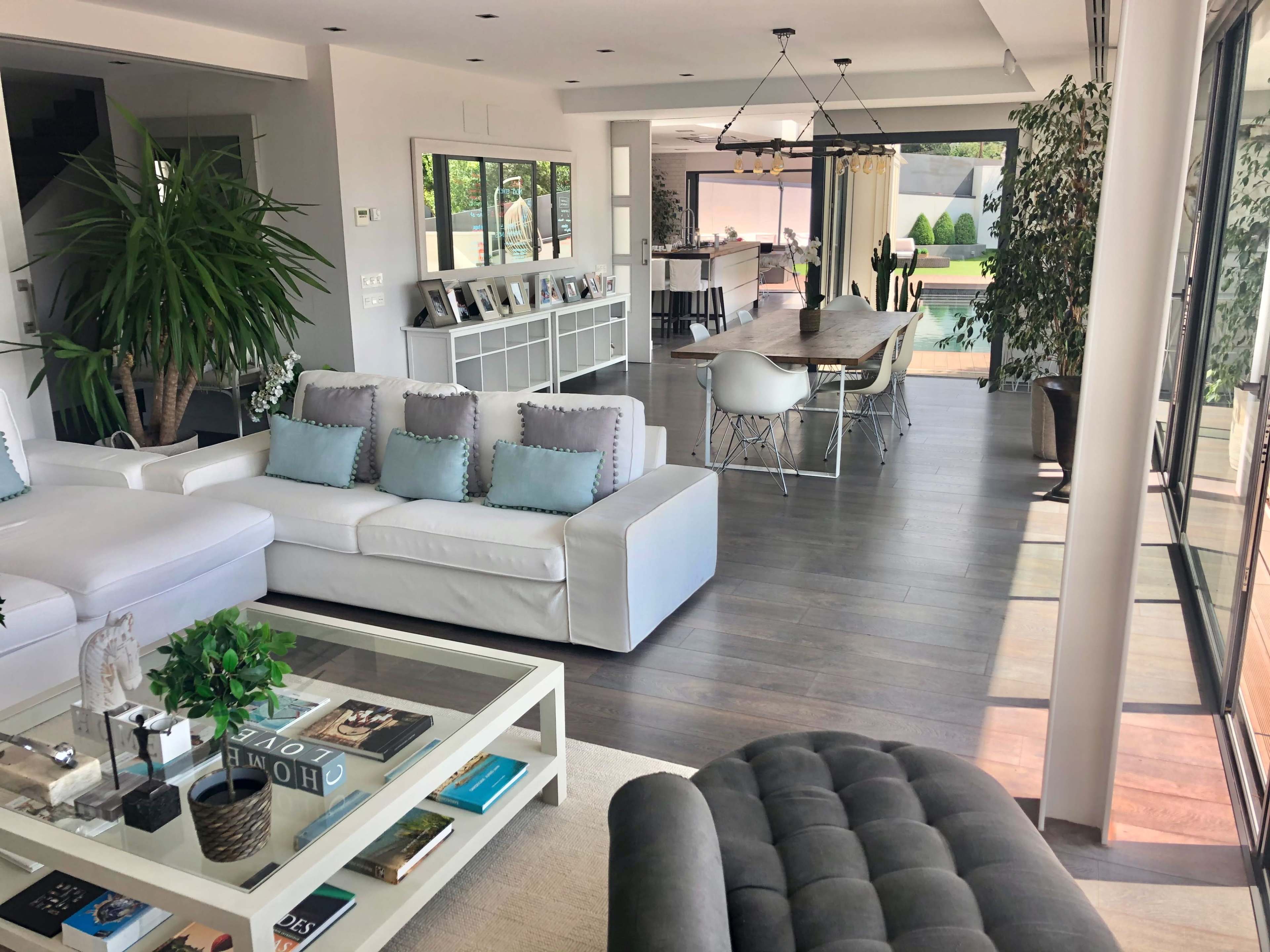 The image shows a modern living space featuring a white sectional couch, a coffee table with books, and a dining area with a large table and chairs, all illuminated by natural light.
