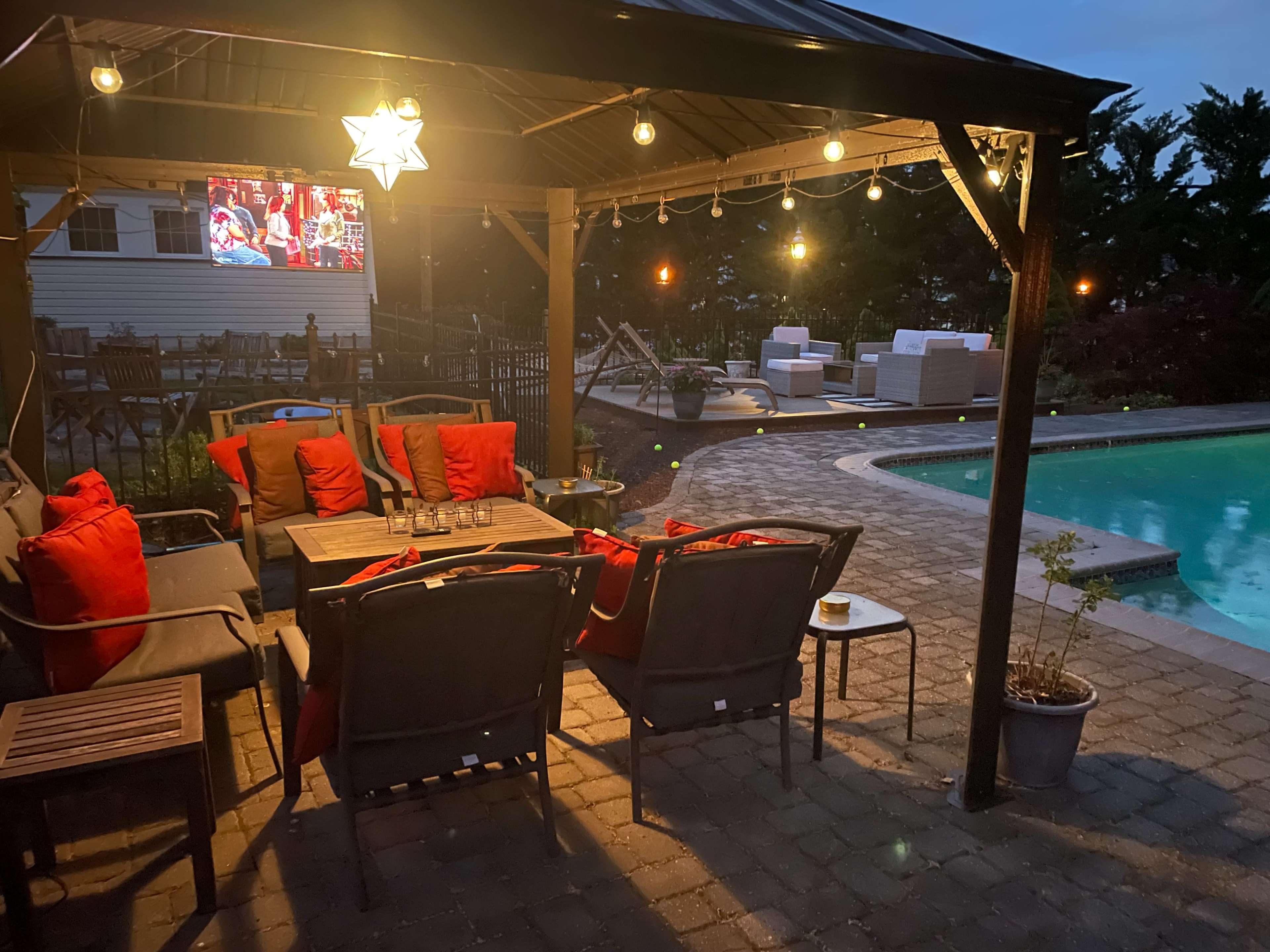 A cozy outdoor seating area under a gazebo features red cushions, a table, and a nearby pool with illuminated landscaping.