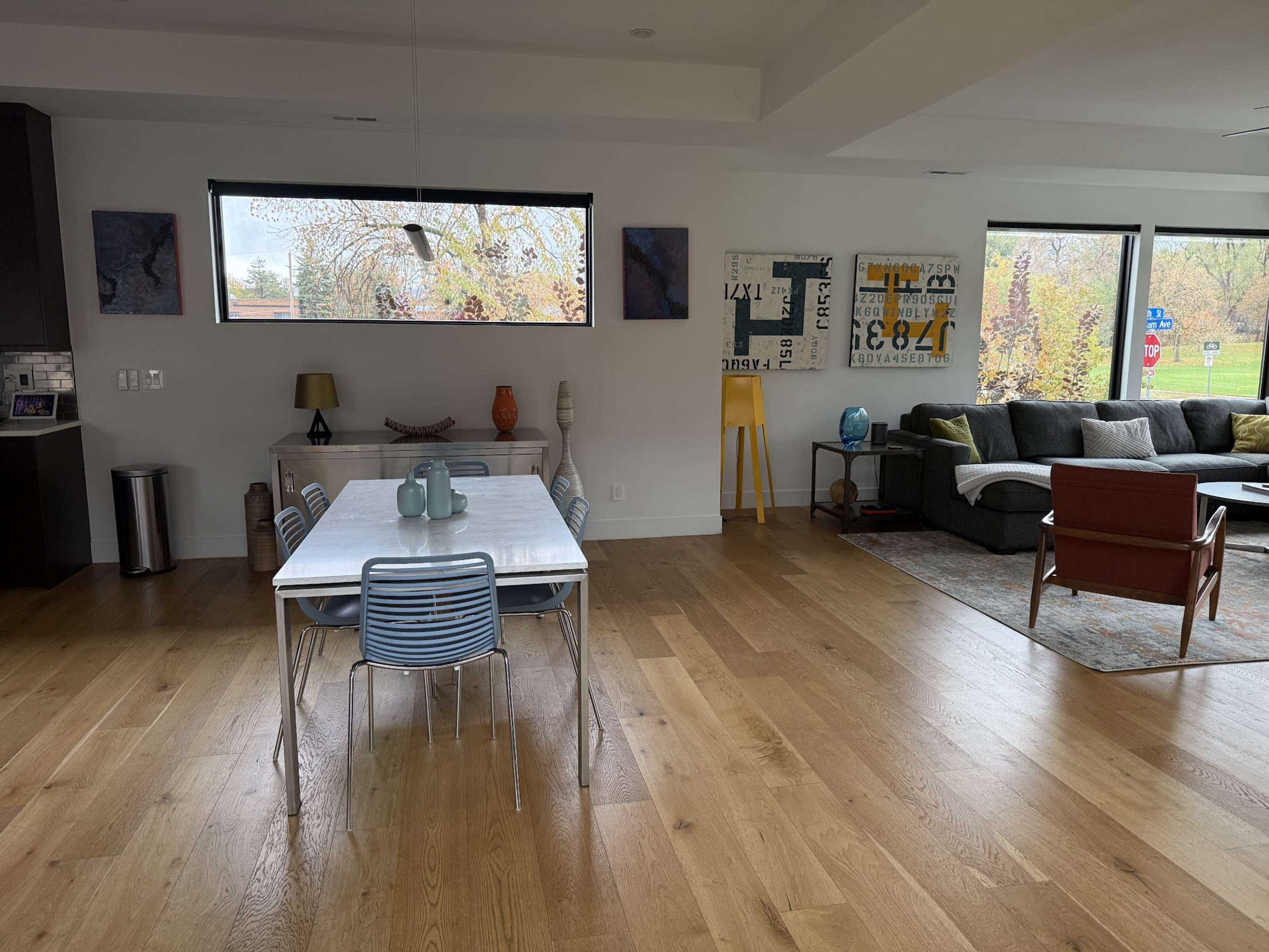 A spacious living area features a dining table with metal chairs, a lounge area with a large sofa, and large windows providing natural light.