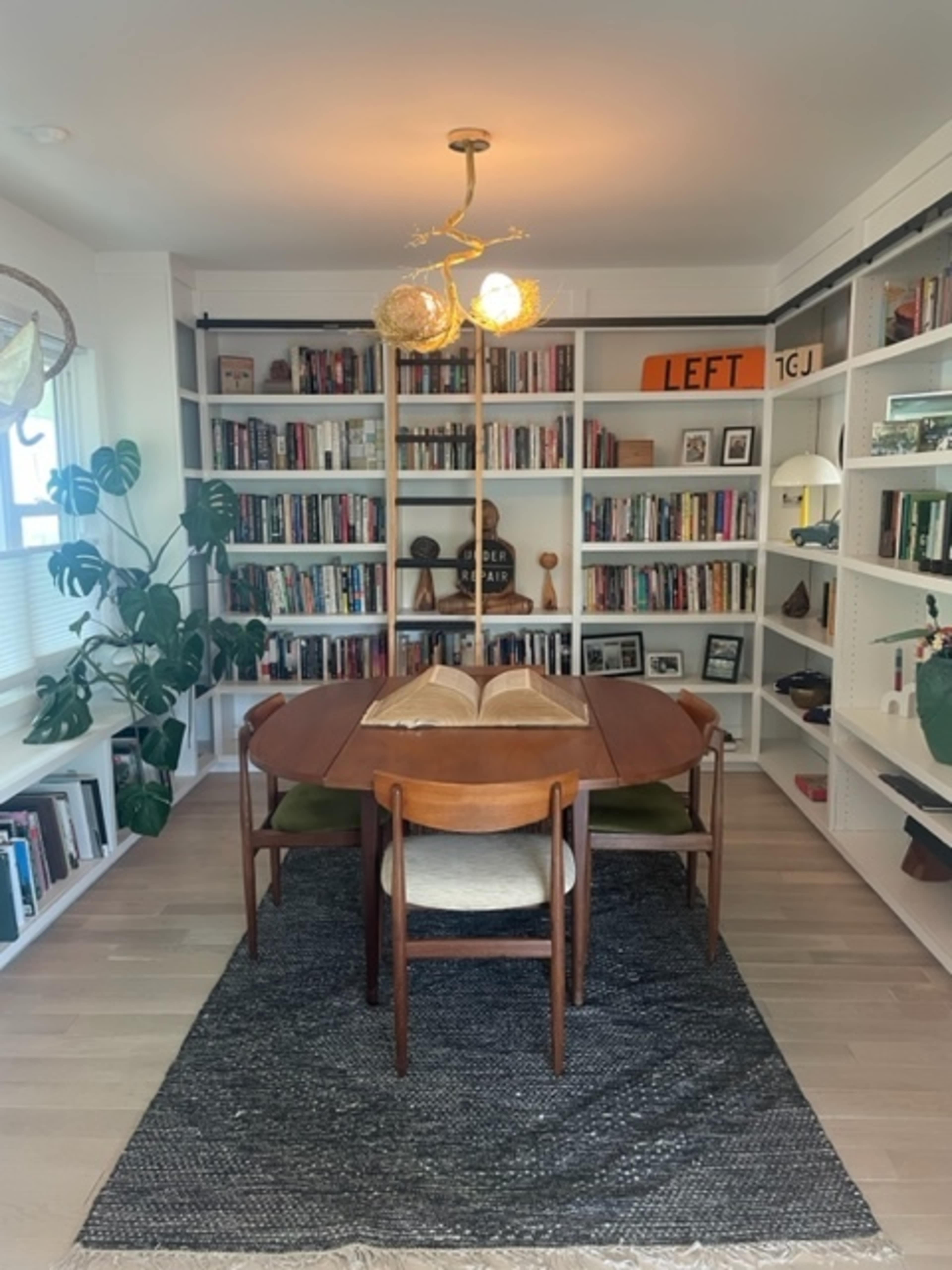 A dining table with wooden chairs is centered in a bright room surrounded by bookshelves filled with various books and decor items.