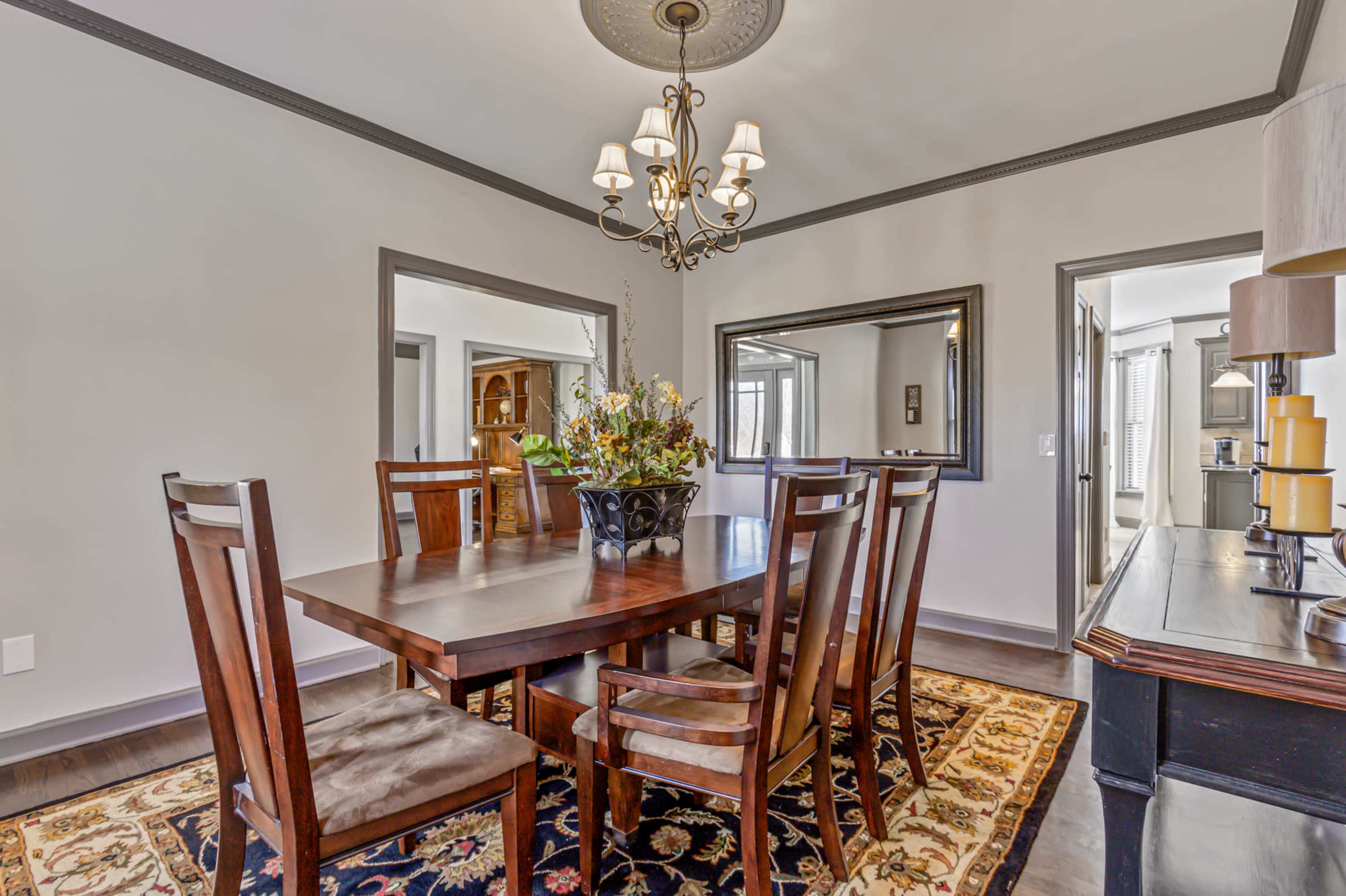 A dining room features a wooden table surrounded by six chairs, a chandelier overhead, and mirrors on the walls.