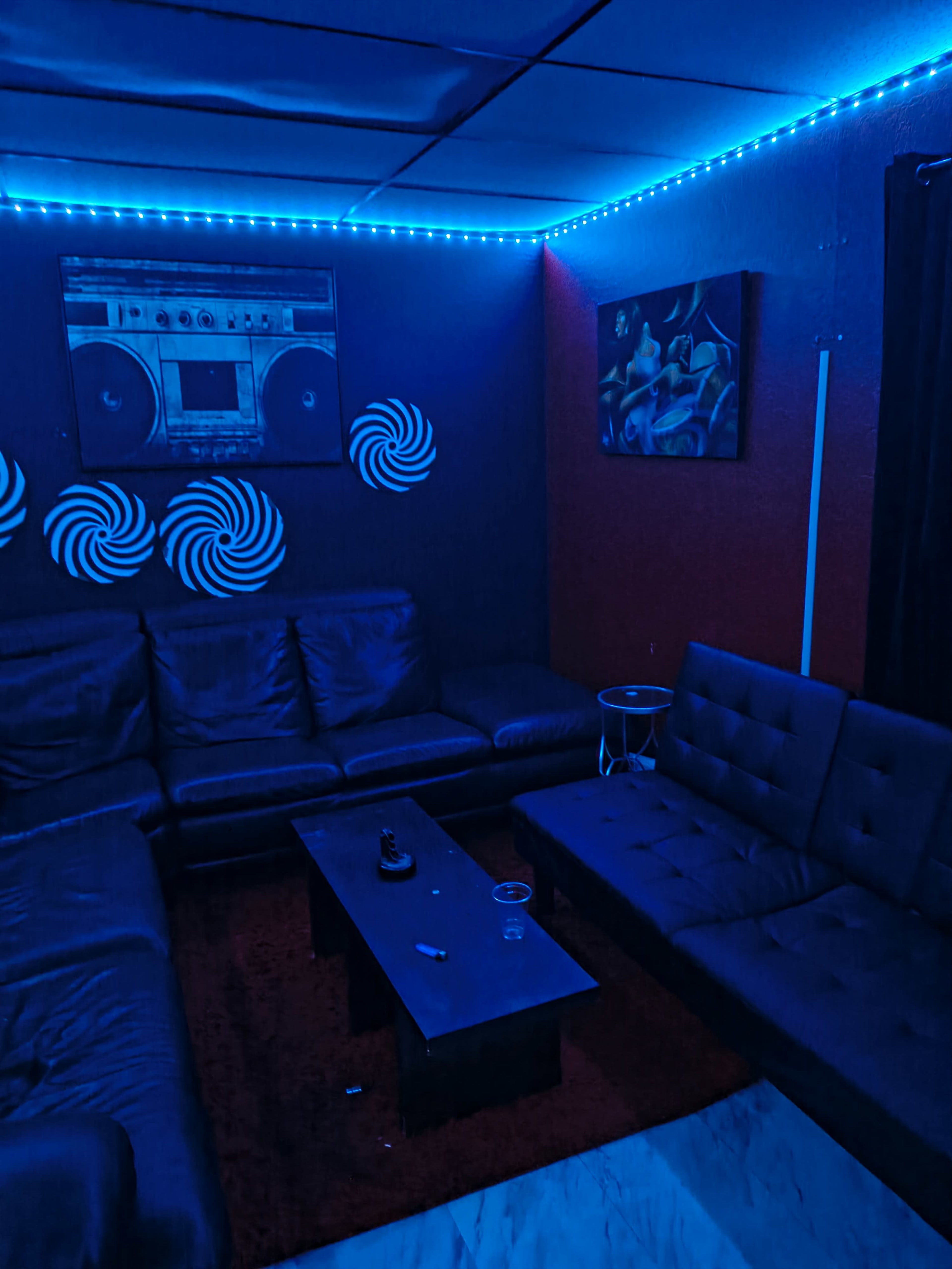 The image shows a dimly lit lounge area with blue LED lights, featuring black leather sofas, a coffee table, and artistic wall decorations.