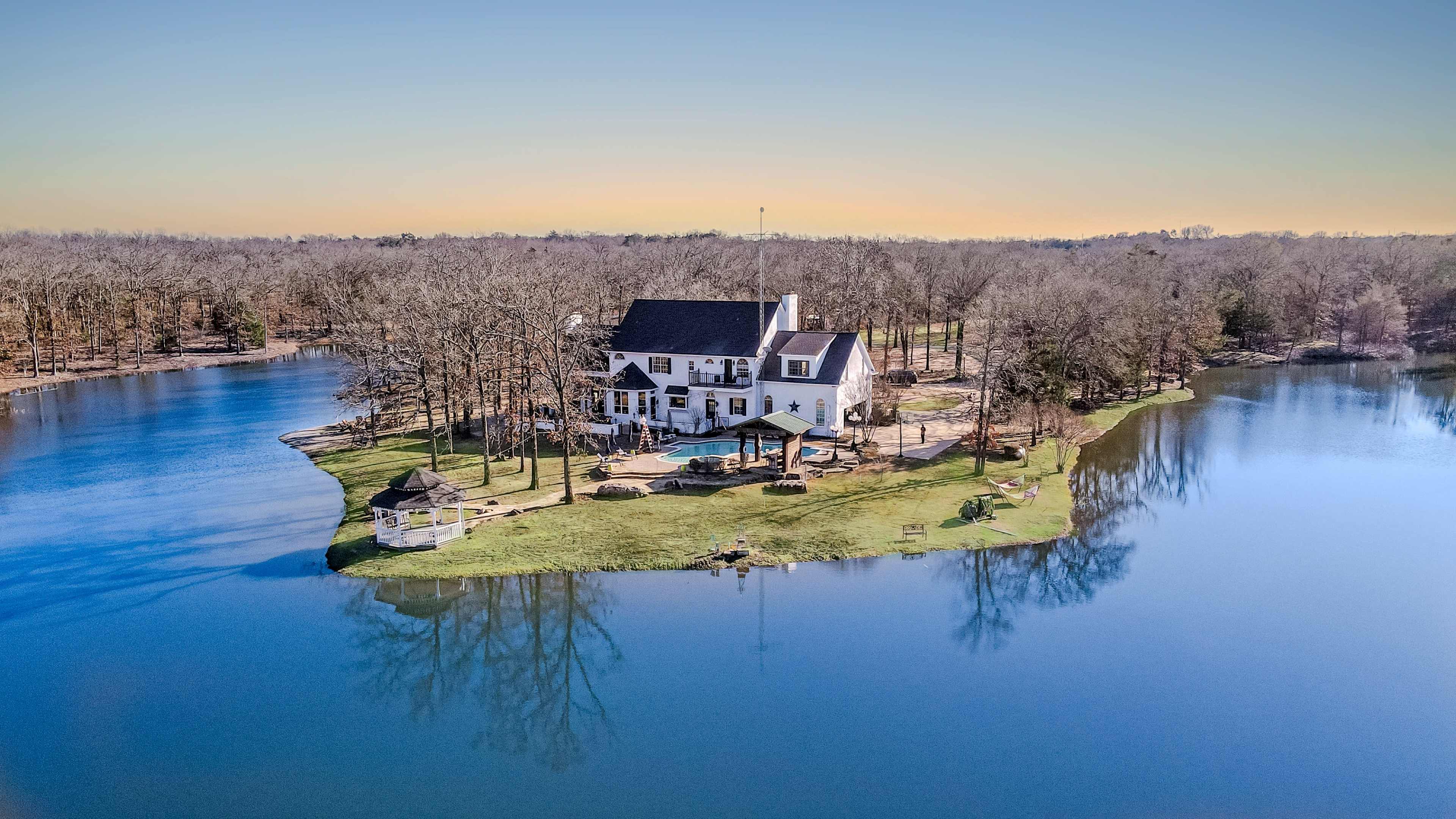 A house is situated on a small island surrounded by calm water and trees in the background.