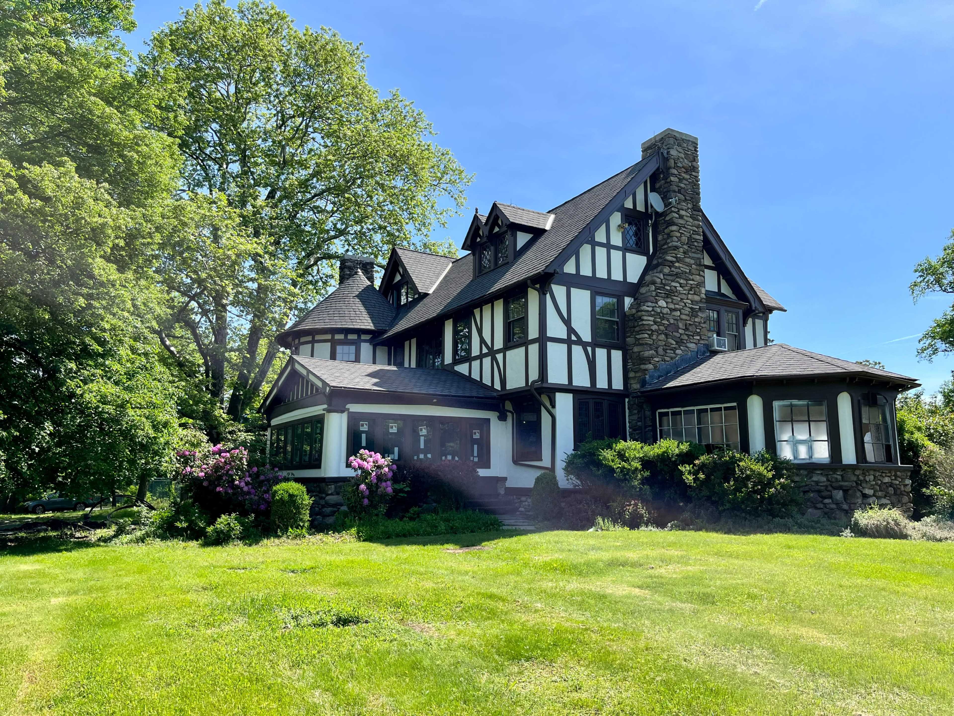 A large, two-story Tudor-style house with stone and timber accents is surrounded by lush green grass and trees.