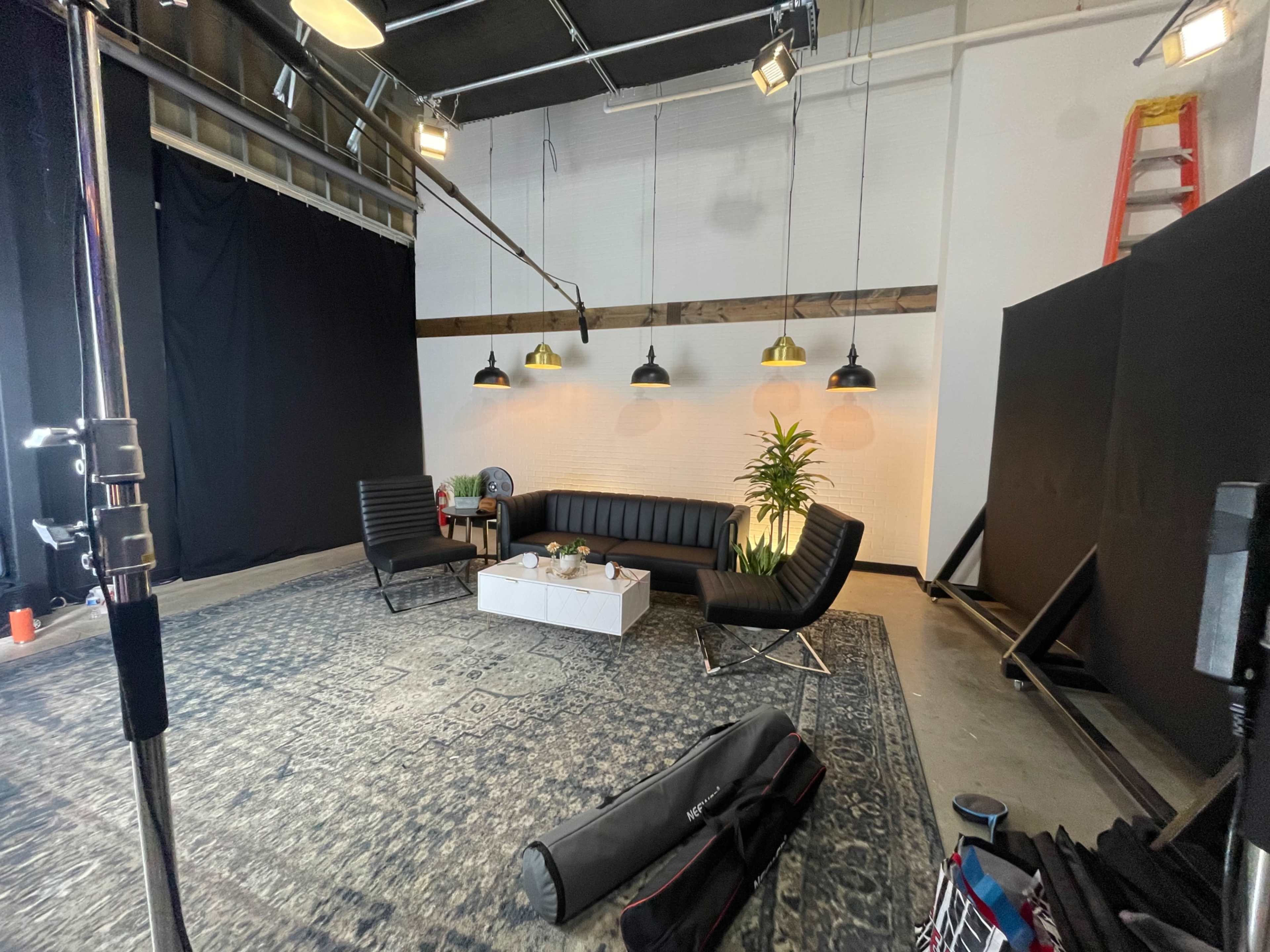 The image shows a studio setup with a black leather sofa, two chairs, a coffee table, a plant, and pendant lights, all arranged on a patterned rug in a well-lit space.
