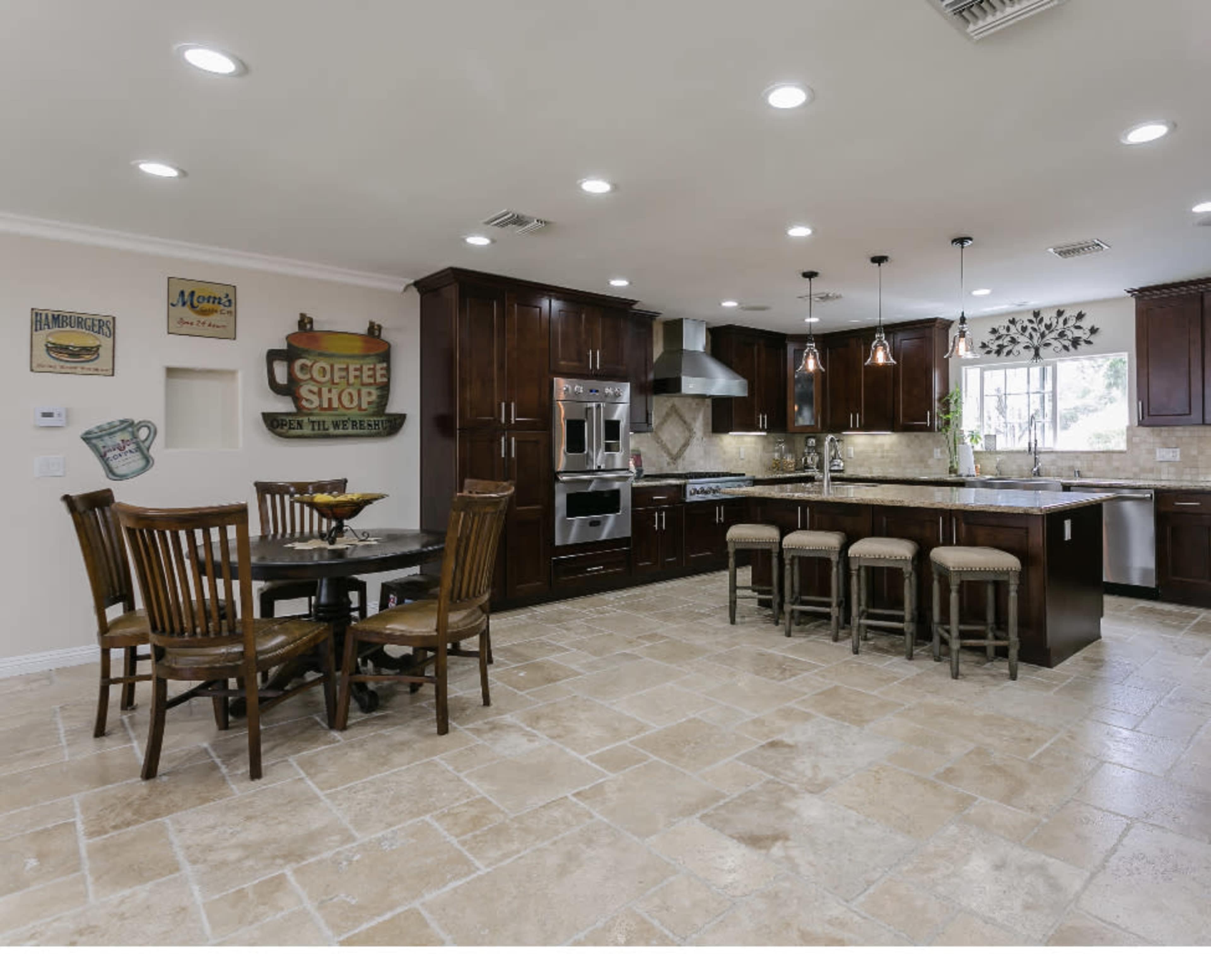 A modern kitchen featuring dark wood cabinetry, a large island with seating, and a dining table set for four, complemented by tiled flooring and decorative wall signs.